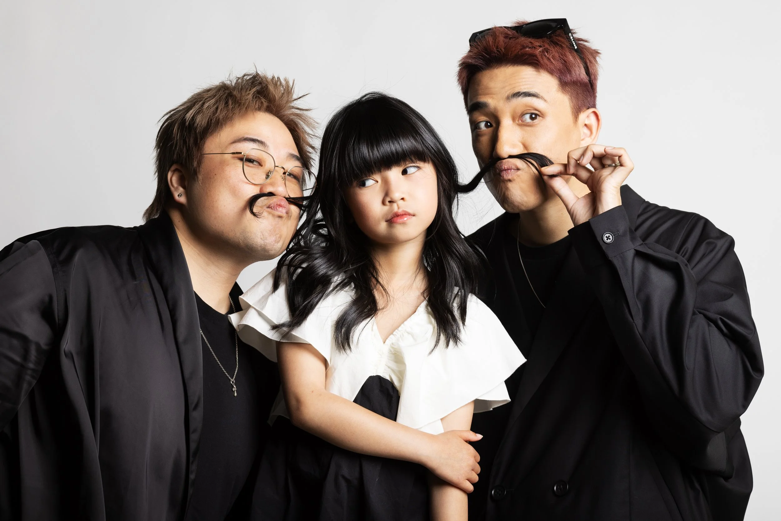 Three young people with fake mustaches, two men and a woman, making playful expressions against a plain background.