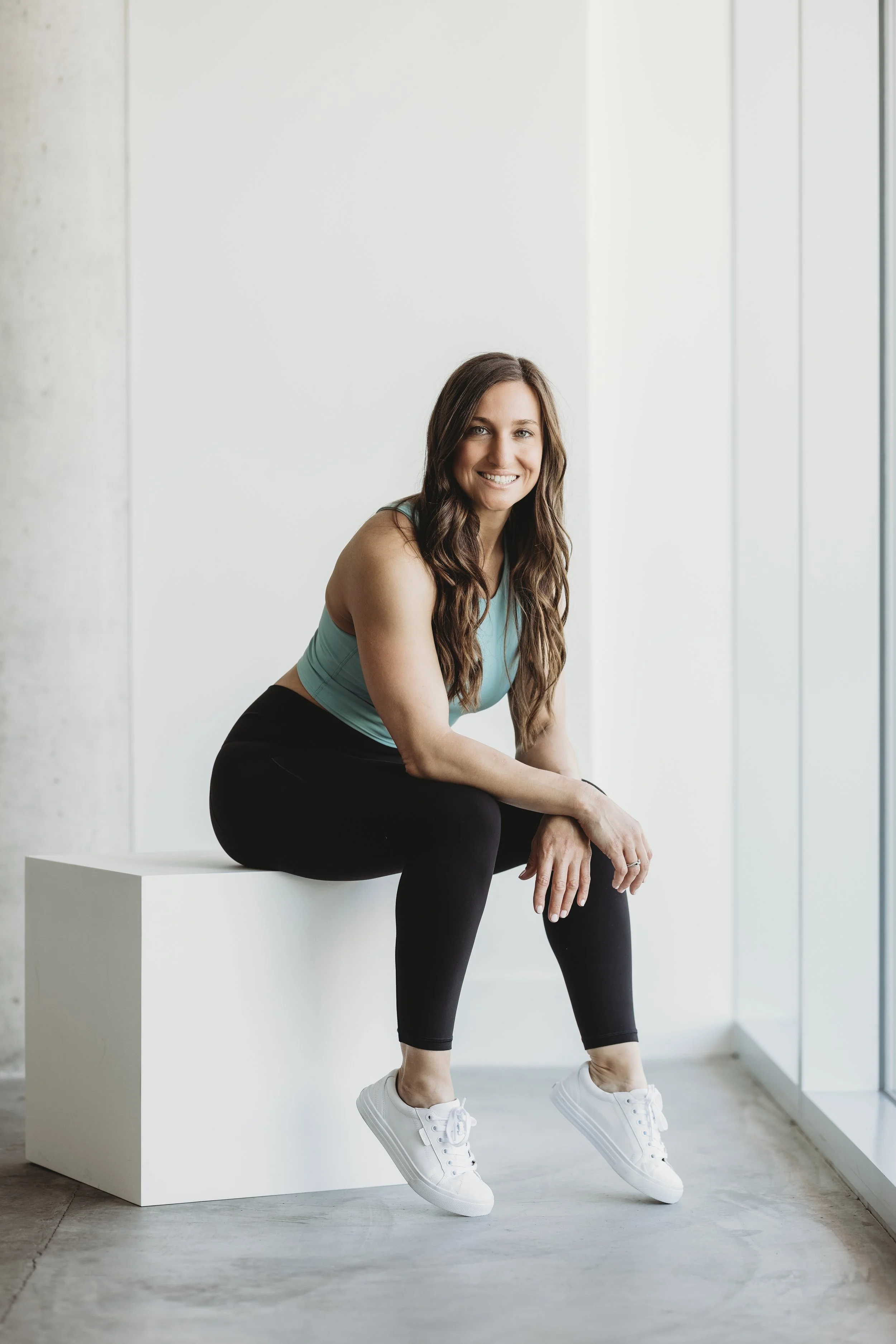 Smiling woman in workout clothes sitting on a white block near a large window in a bright, modern space.