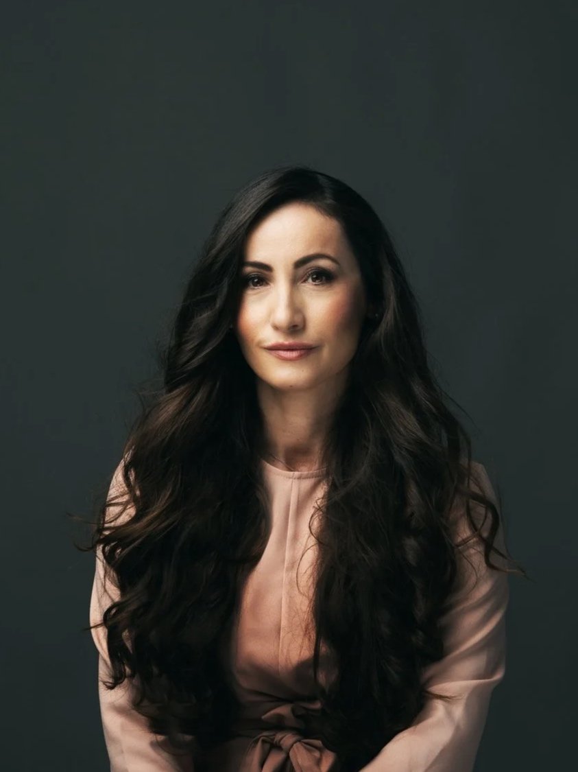 Portrait of a woman with long, dark, wavy hair, wearing a light-colored top, against a dark gray background.