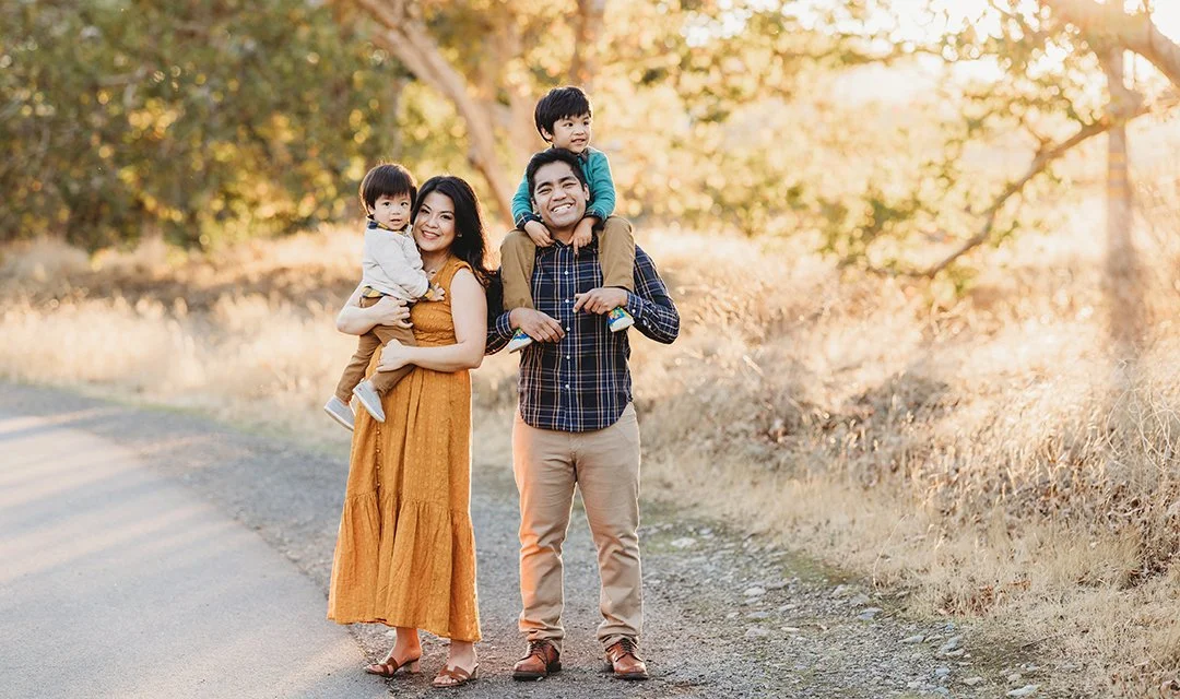 Family of four walking outdoors on a dirt path, with trees and golden sunlight in the background, enjoying a sunny day.