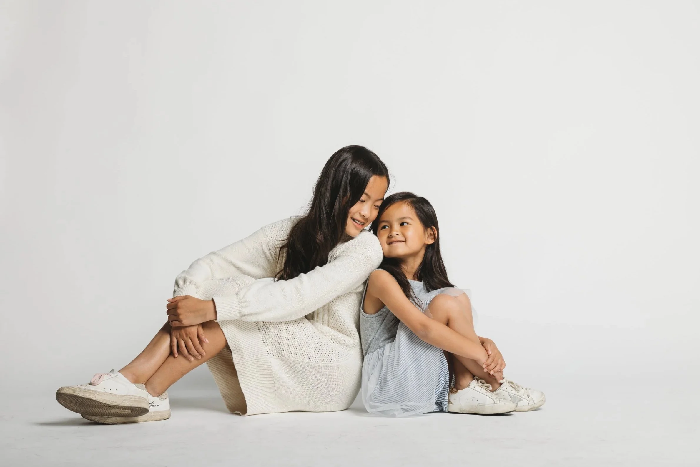 A woman and a young girl sitting on the floor, hugging and smiling at each other, against a plain white background.