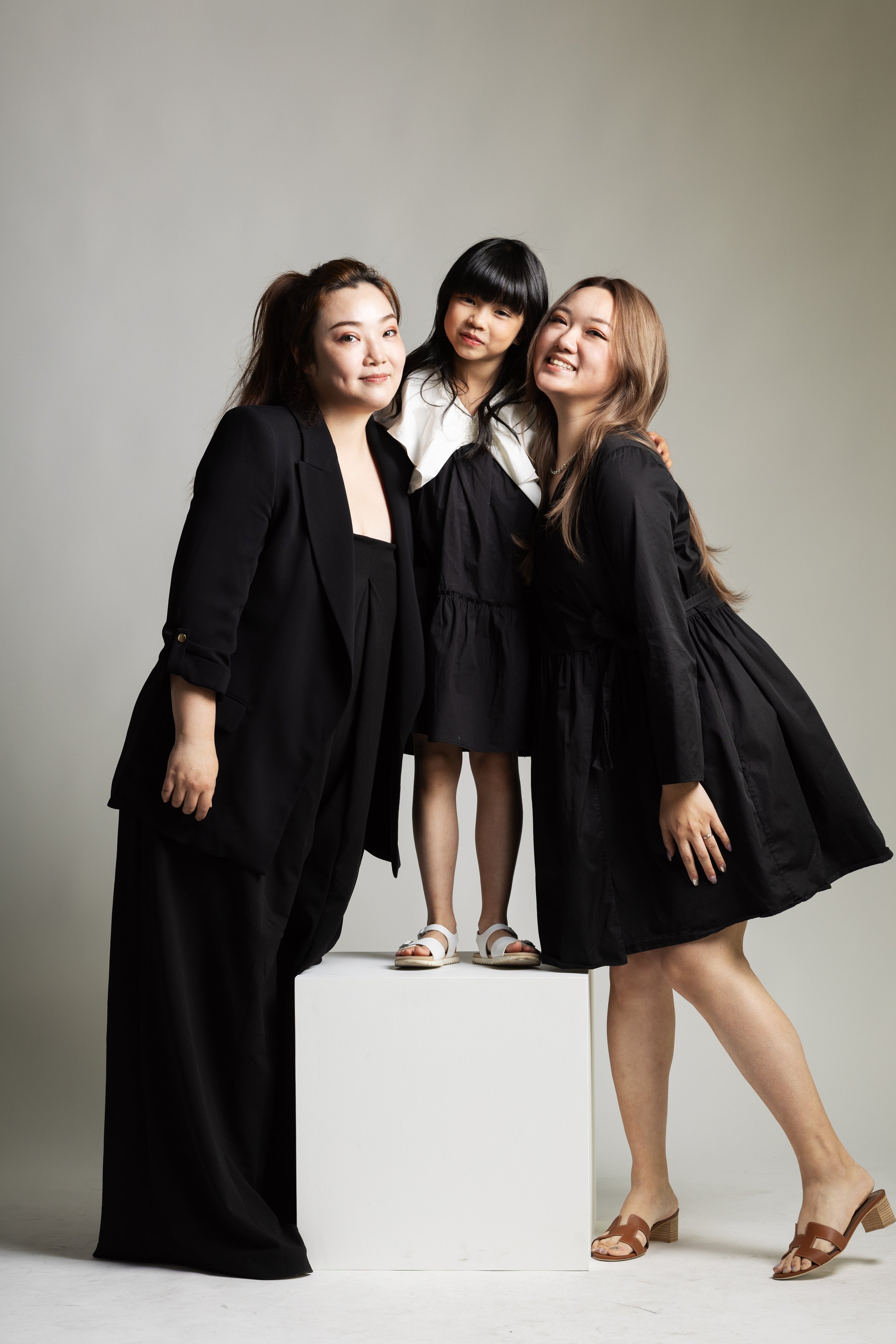 Three women, two adults and one child, standing around a white pedestal, smiling for a portrait in a neutral studio setting. The adults wear black outfits; the child wears a black dress with a white collar and white sandals.