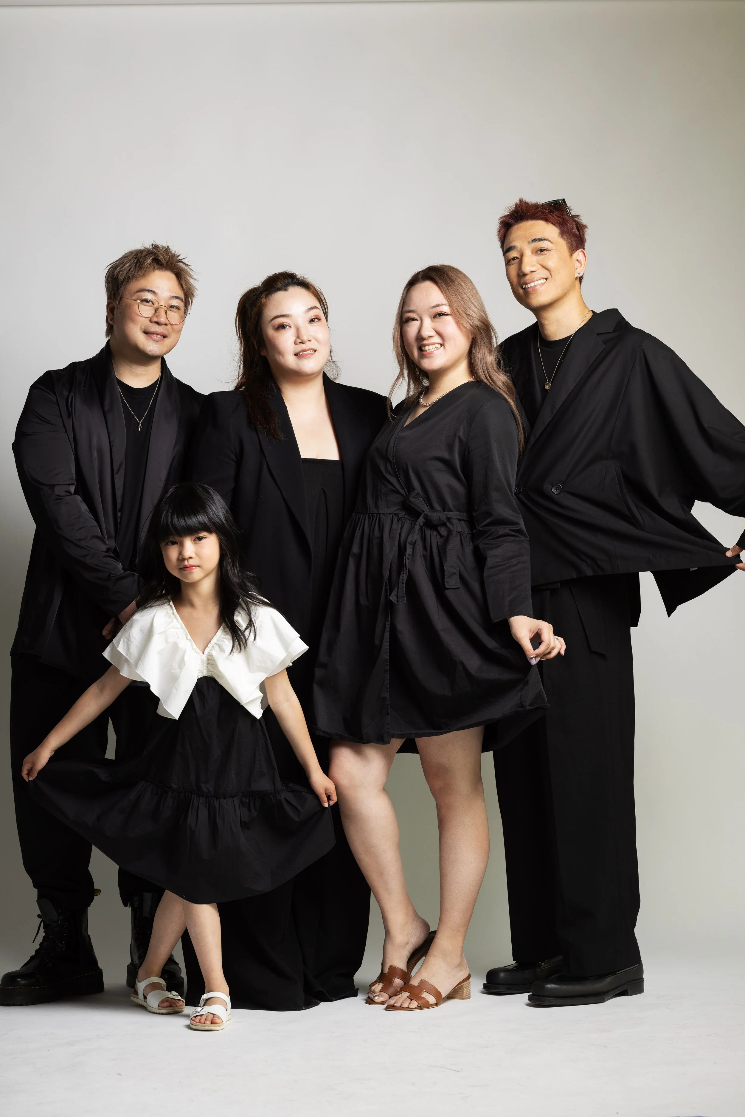 Group of five people, four adults and one child, standing together in black and white outfits, posing against a plain white wall.