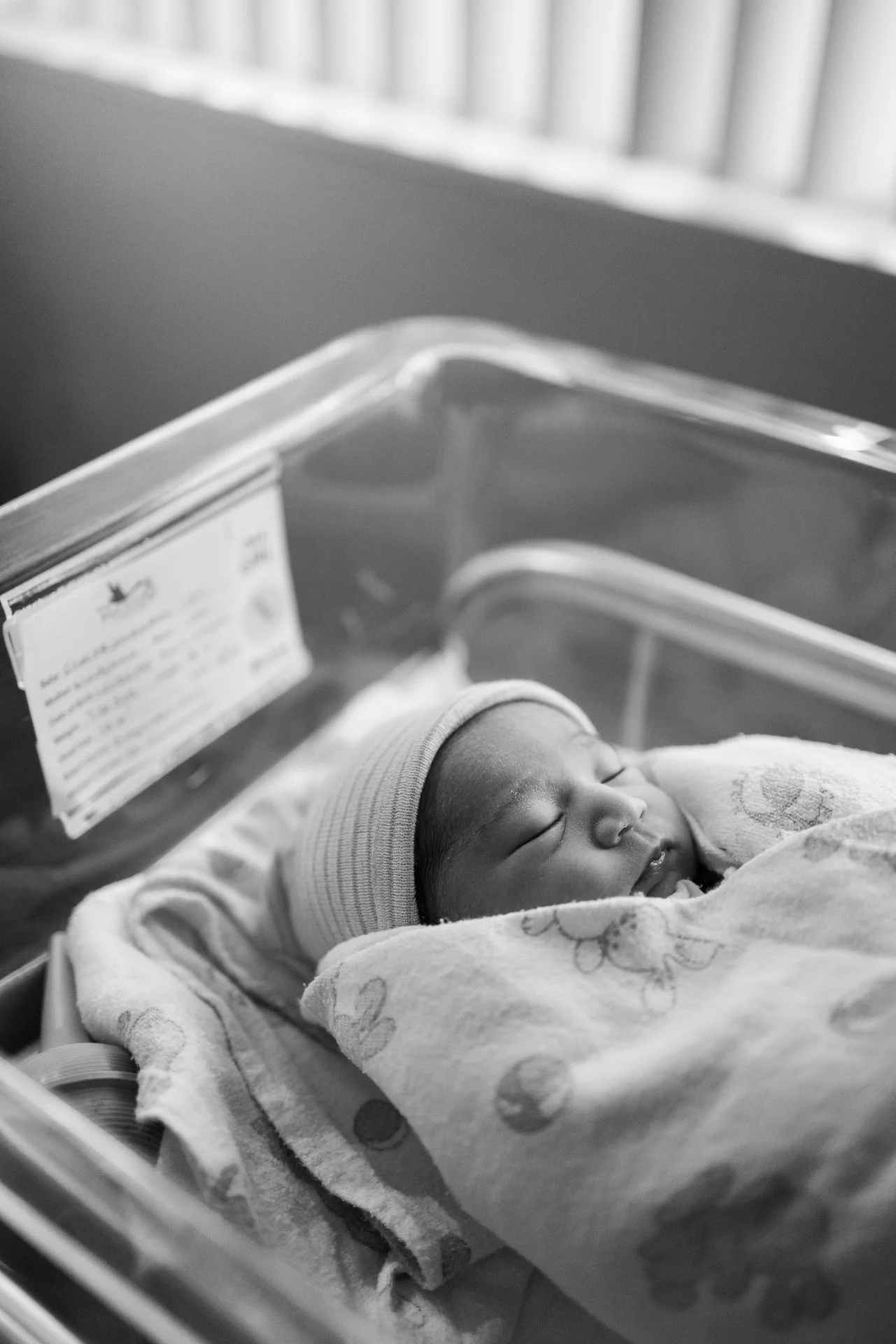 A newborn baby sleeping in a hospital bassinet, wrapped in a blanket and wearing a hat.
