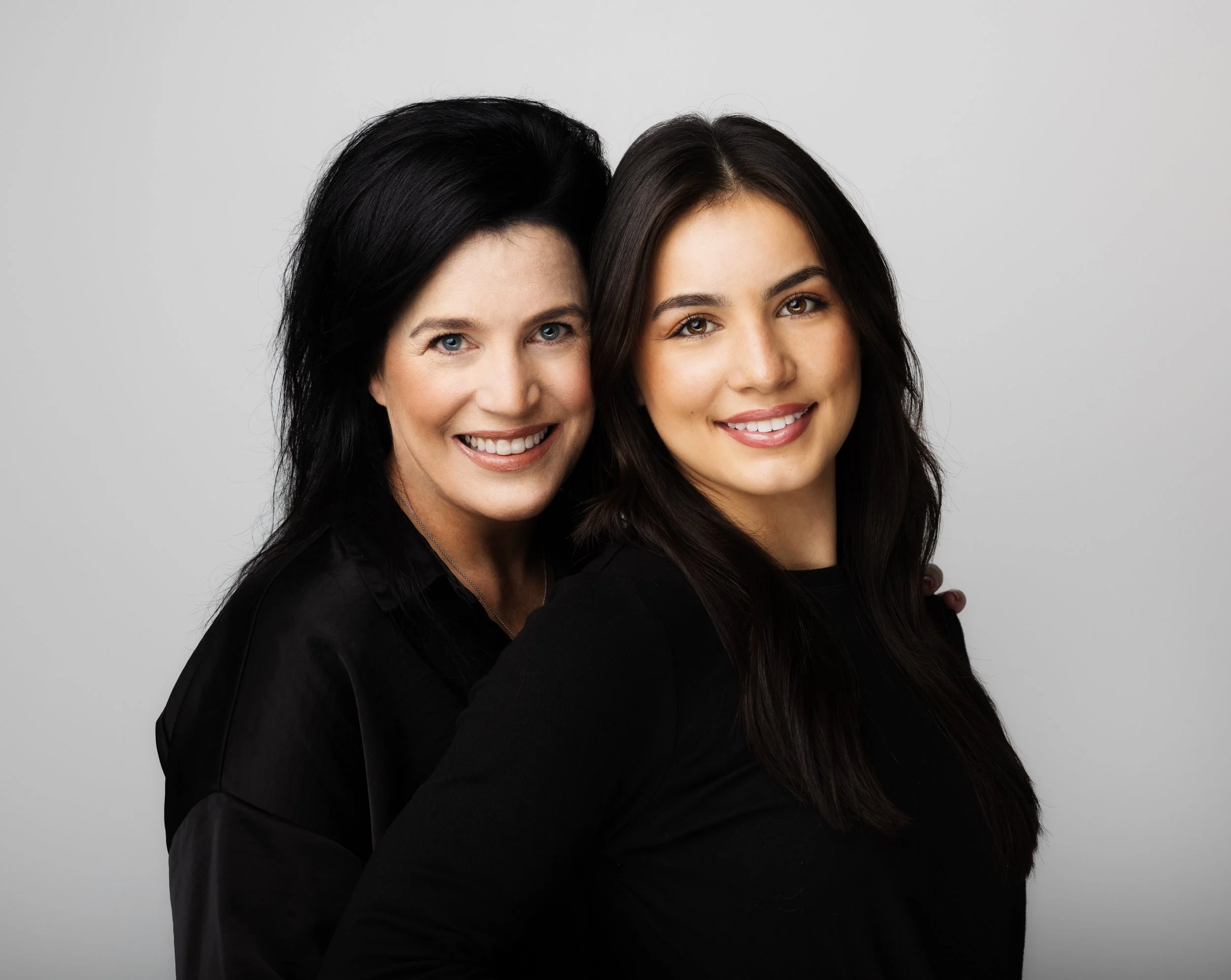 Two women smiling and embracing each other against a plain gray background.
