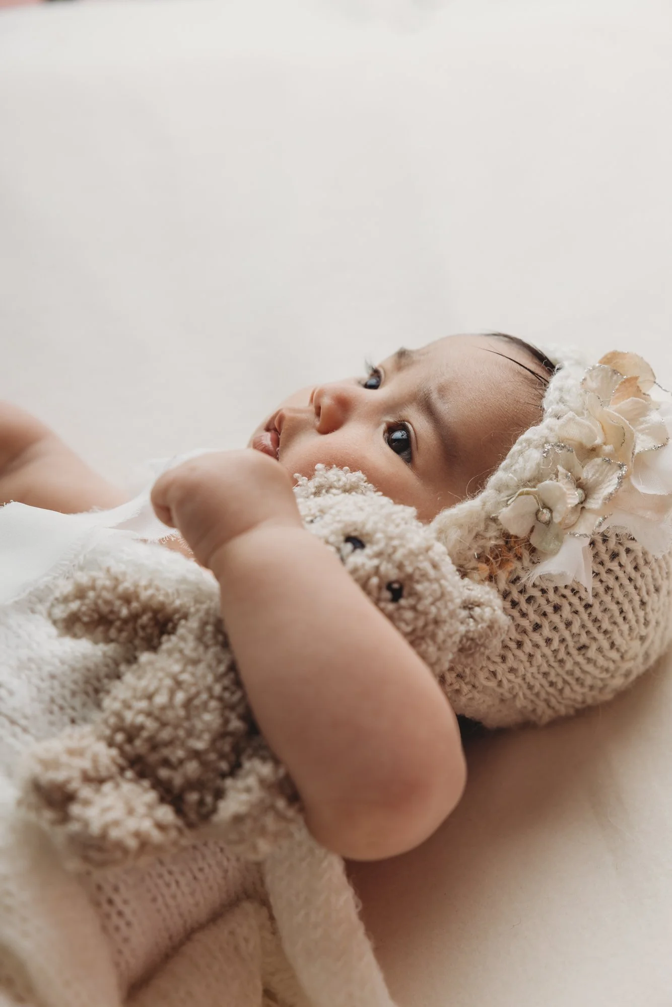 Close-up of a baby lying on a soft surface, wearing a white bonnet decorated with flowers and pearls, wrapped in a knitted blanket, looking up and to the side.