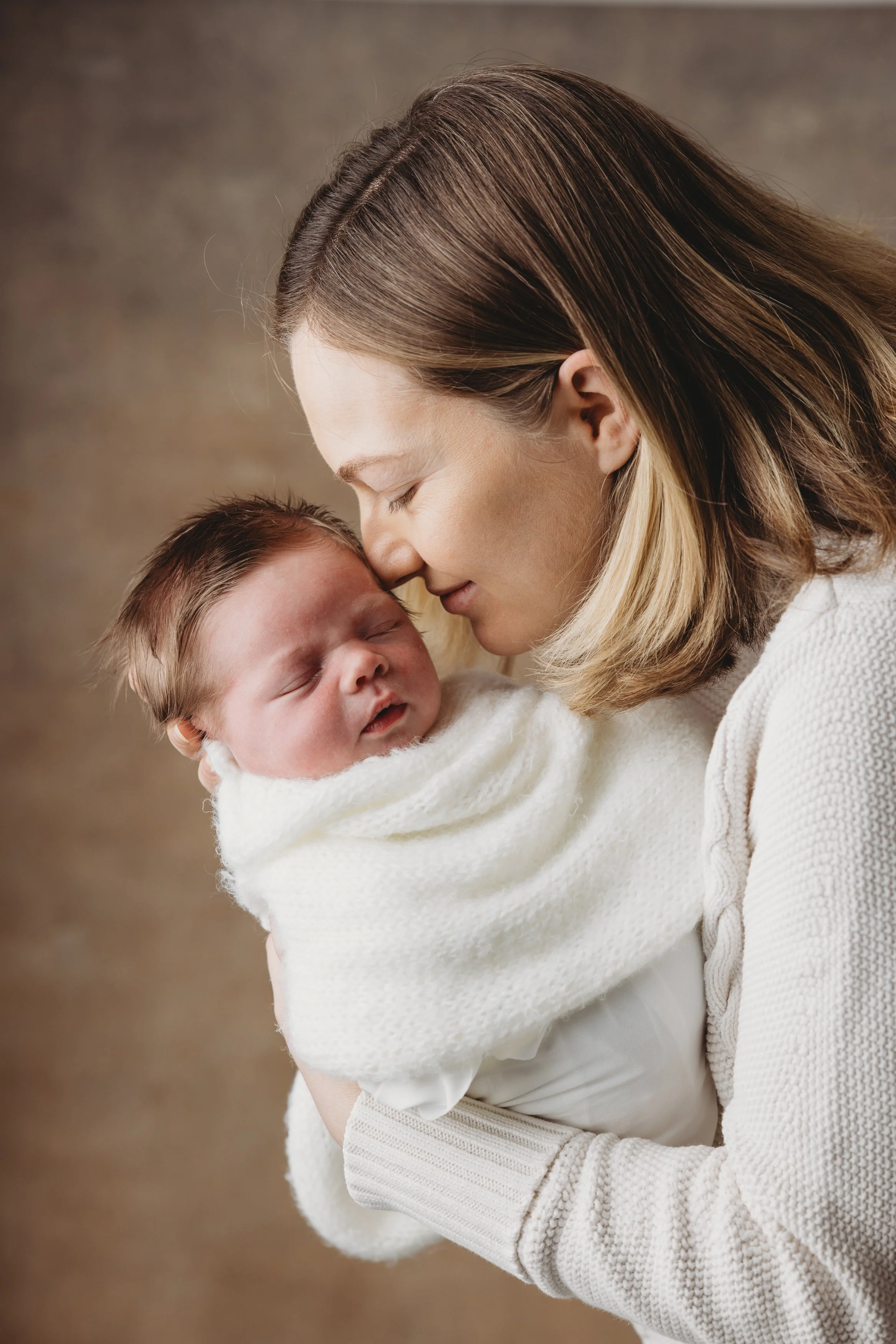 A woman with shoulder-length brown hair lovingly holding a sleeping baby wrapped in a white blanket.