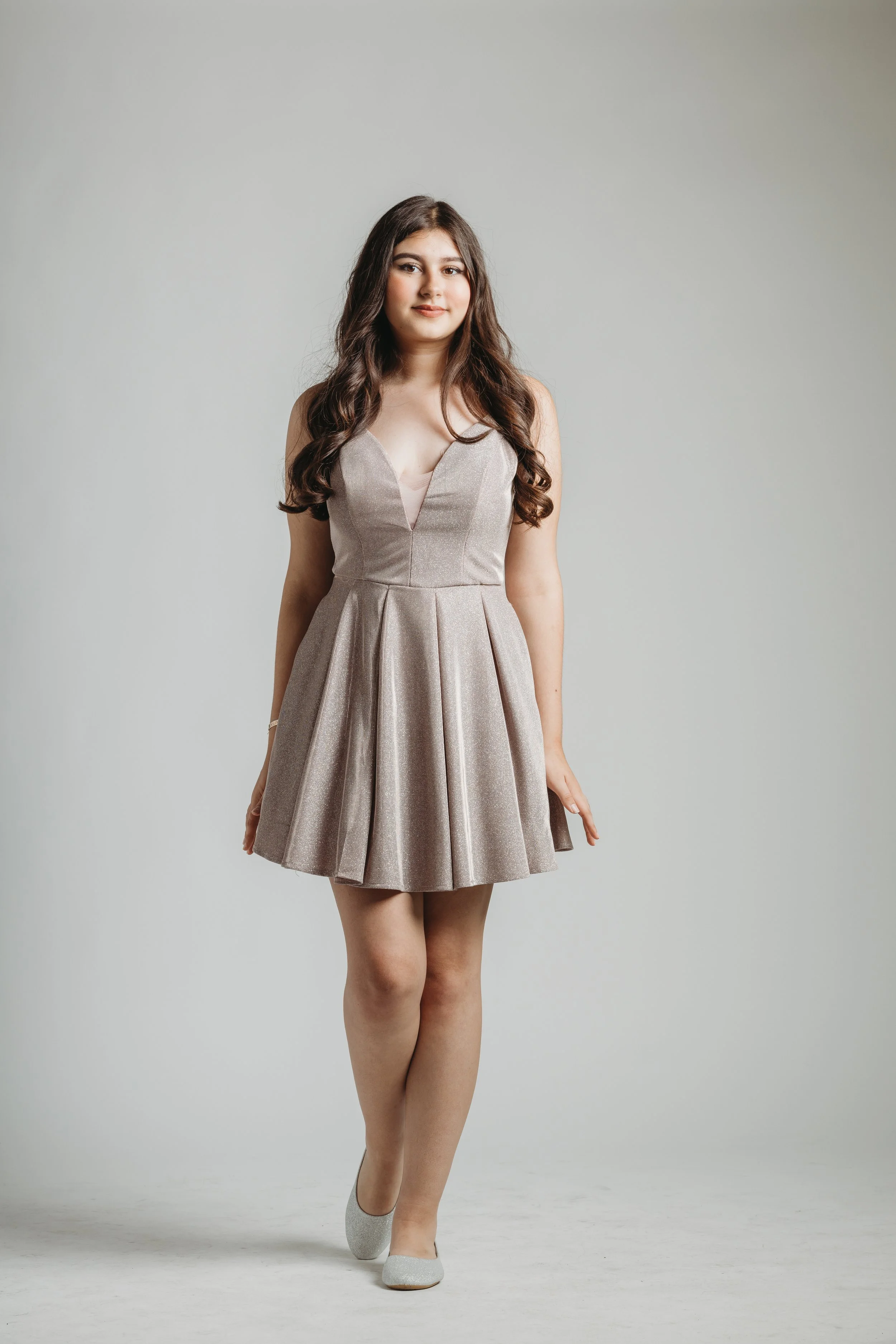 A young woman with long, wavy brown hair, wearing a light-colored, knee-length dress with a V-neckline and pleated skirt, and silver shoes, standing against a plain light grey background.