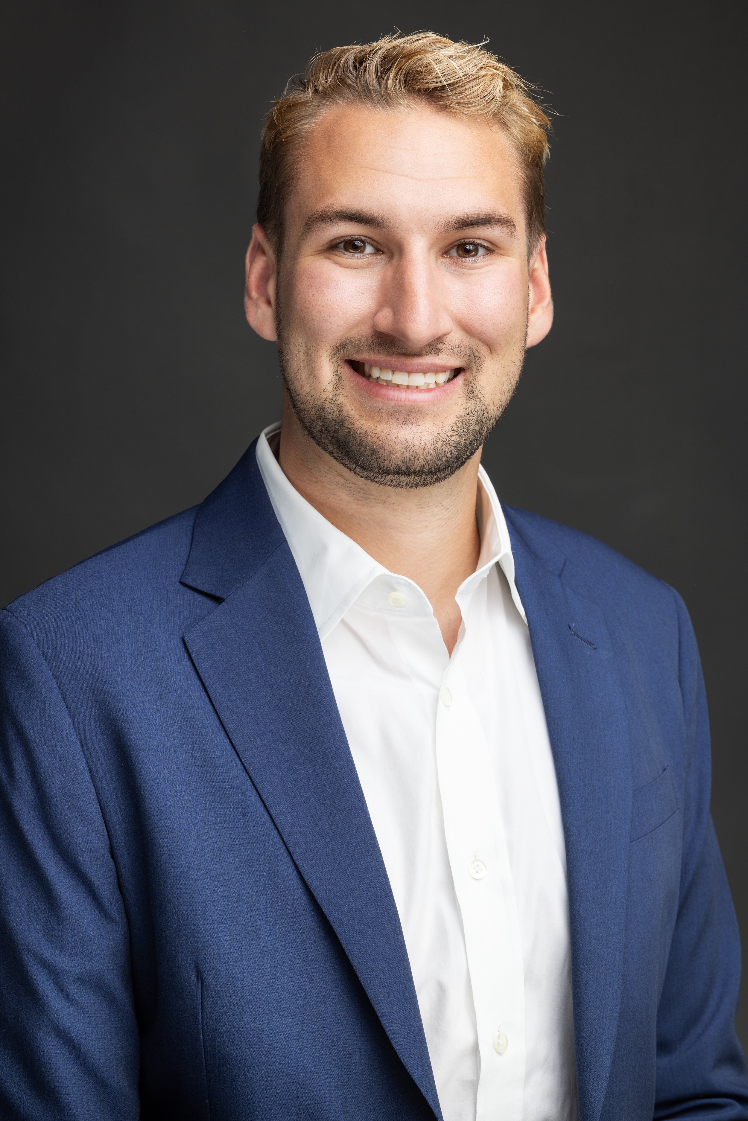 Professional headshot of a young man with light brown hair, smiling, wearing a white collared shirt and a blue blazer, against a dark gray background.