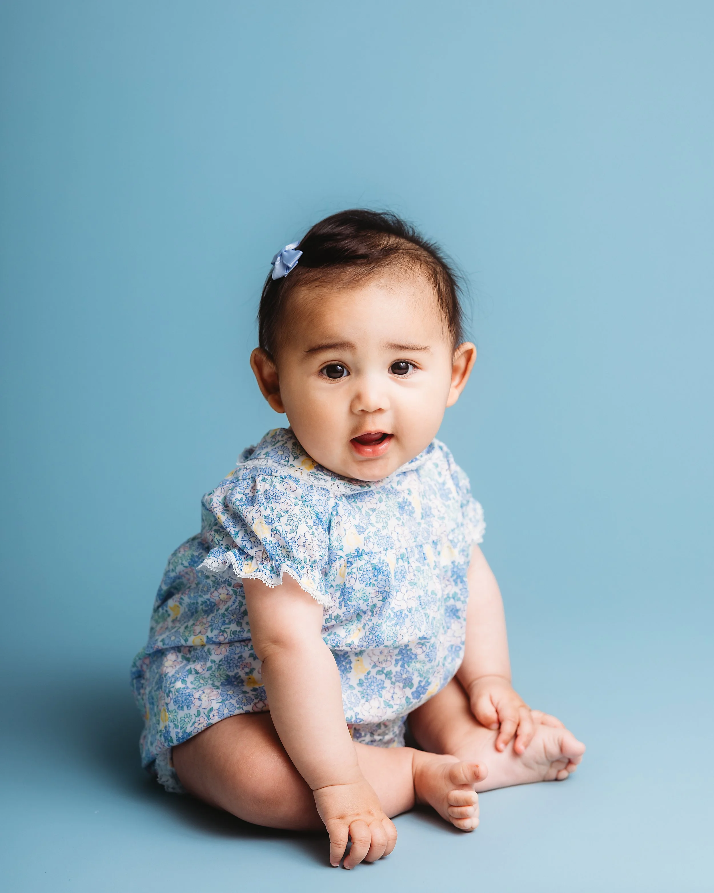 A baby girl sitting on the floor against a blue background, wearing a floral dress and a blue hair clip, looking at the camera with a curious expression.