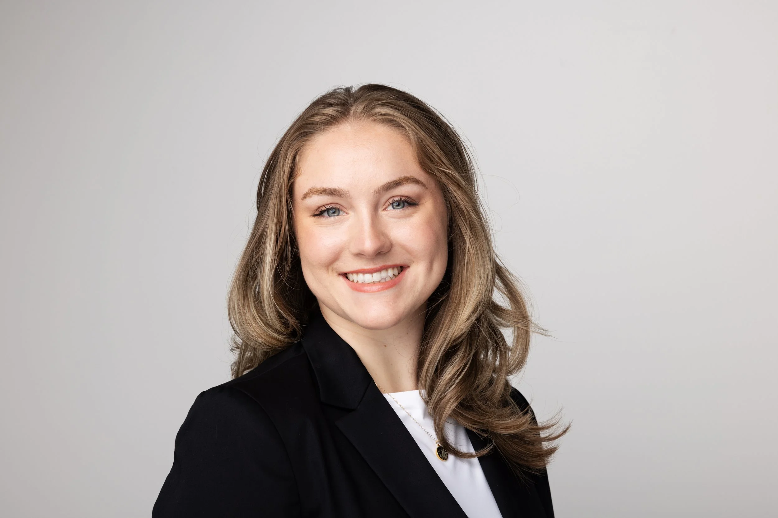 A young woman with long wavy blonde hair, blue eyes, and a bright smile, wearing a black blazer over a white shirt, standing against a plain light gray background.