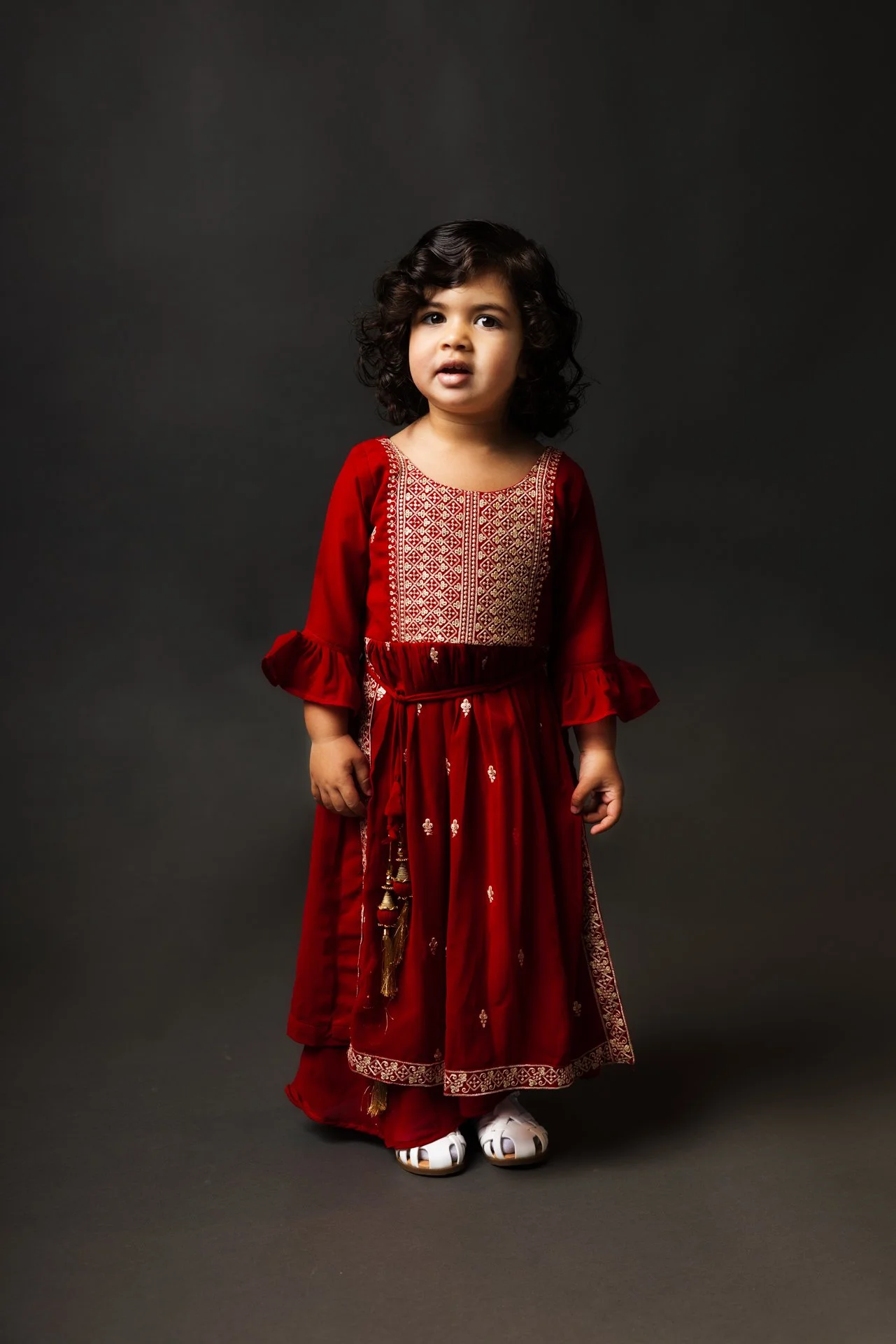 Young girl with curly hair wearing a traditional red embroidered dress and white sandals, standing against a dark gray background.