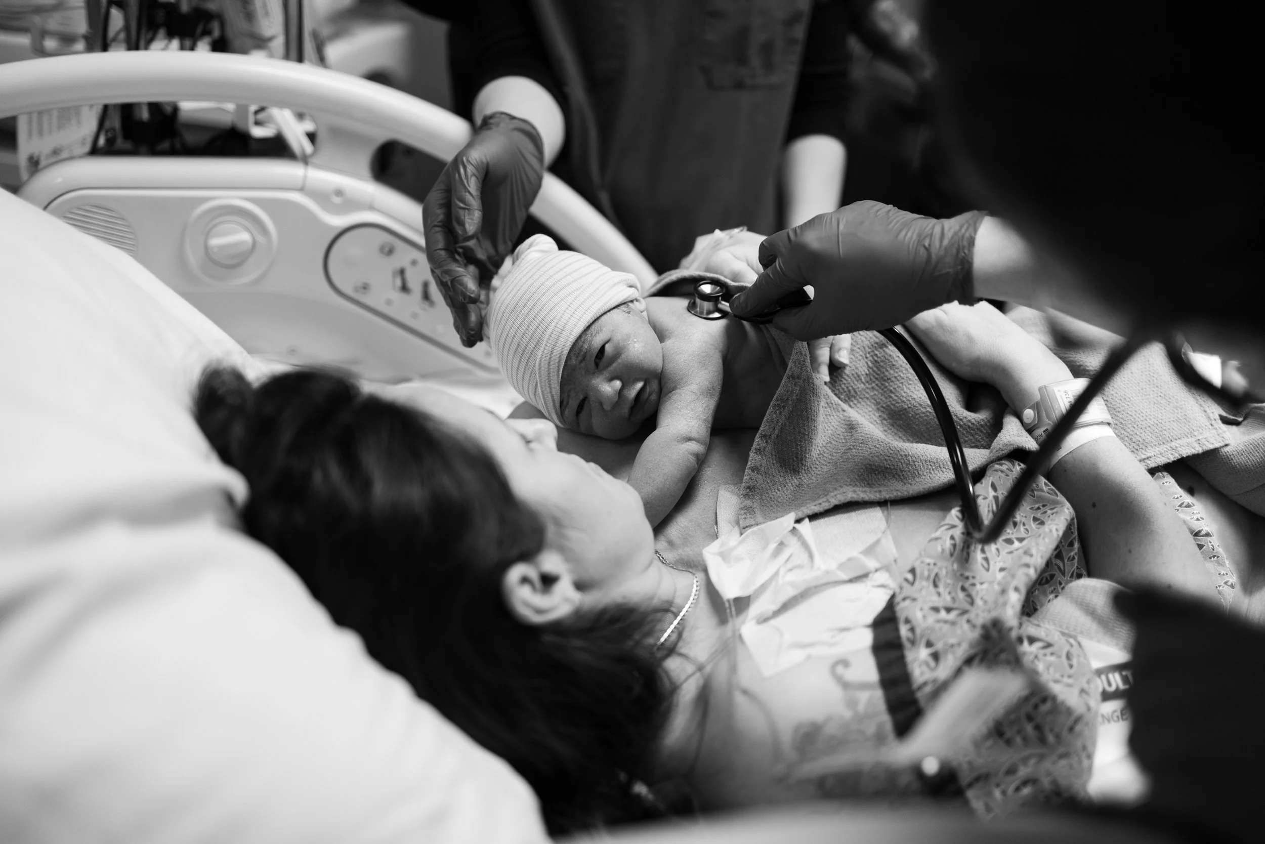 A newborn baby in a hospital bed, receiving medical attention with a stethoscope, surrounded by healthcare staff wearing gloves, in a clinical setting.