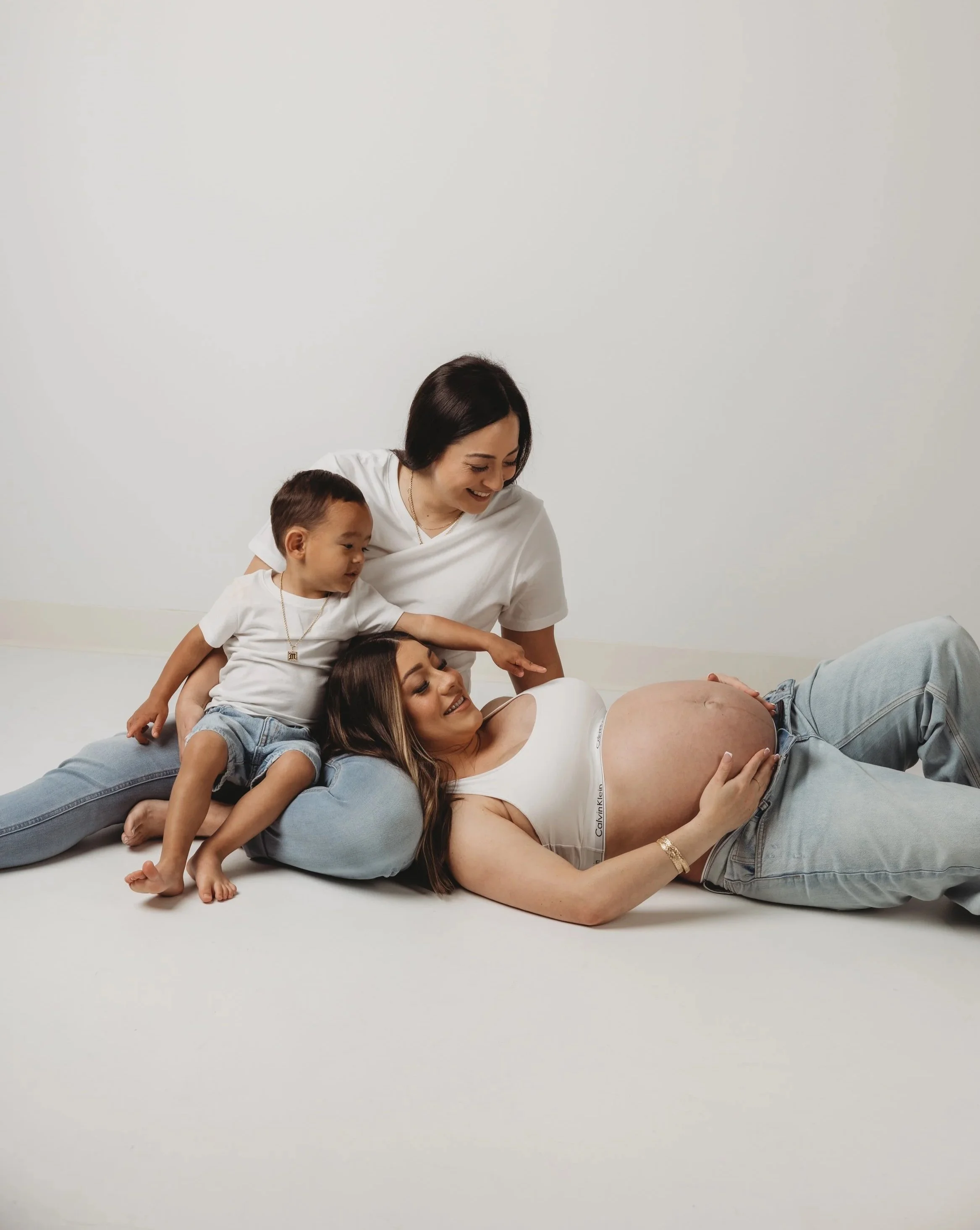 A pregnant woman lying on the floor with her head on her arm, surrounded by three children, all smiling and touching her belly, in a minimal white space.