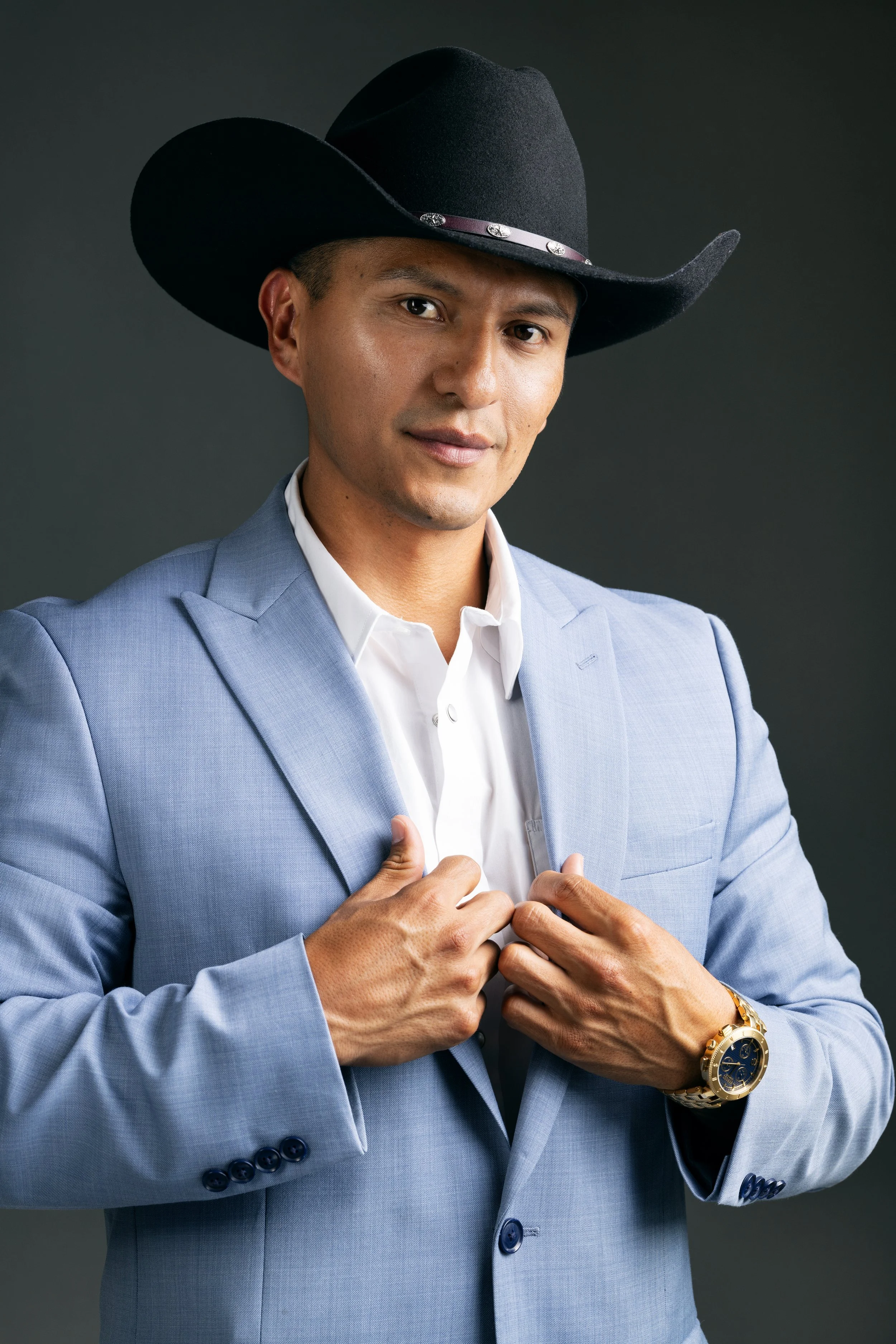 A man wearing a light blue suit, white shirt, a black cowboy hat with silver accents, and a gold watch on his left wrist, standing against a dark background.