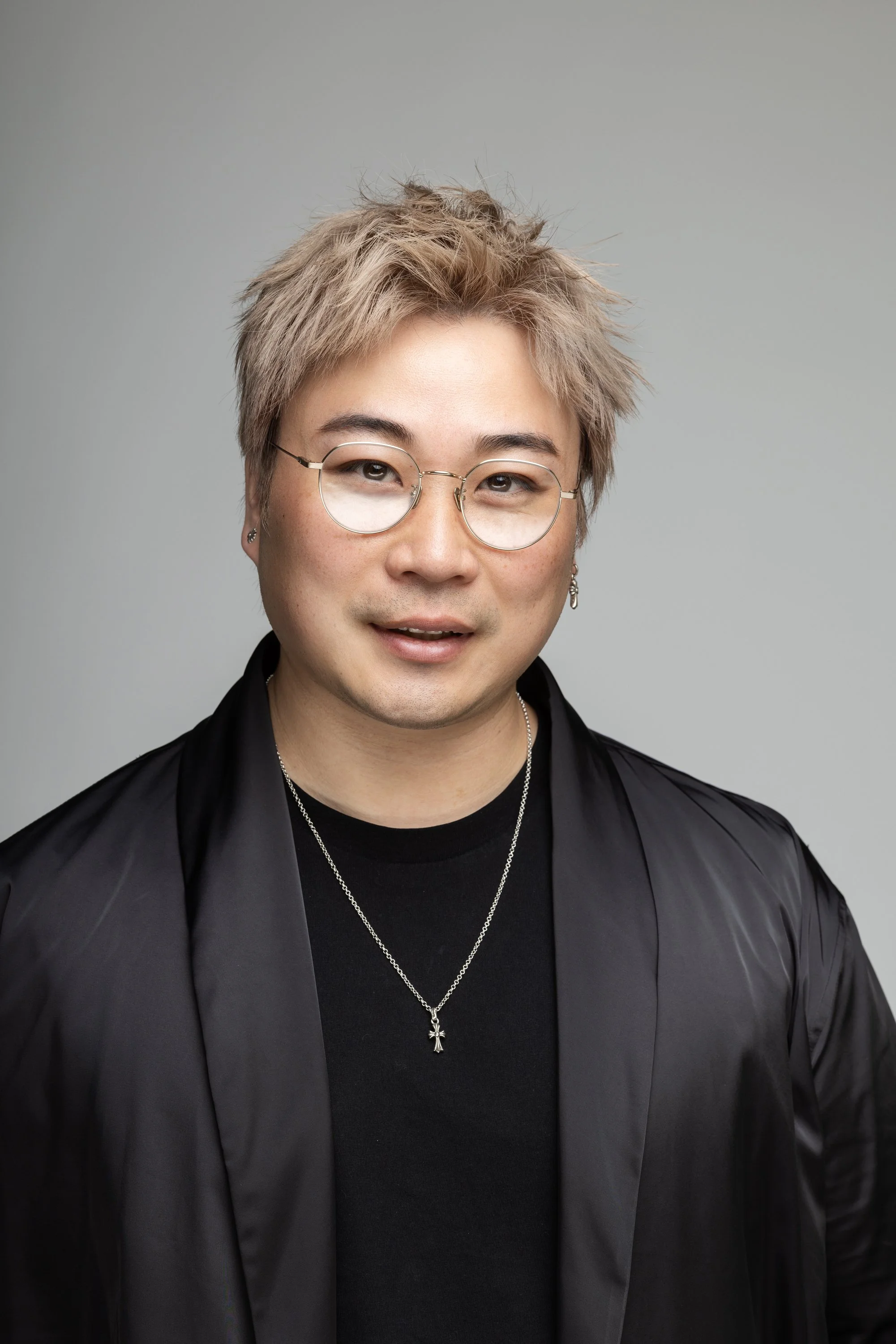 Portrait of a young person with short messy blond hair, wearing clear glasses, a black jacket, black t-shirt, and a silver necklace with a cross pendant, smiling against a plain light gray background.