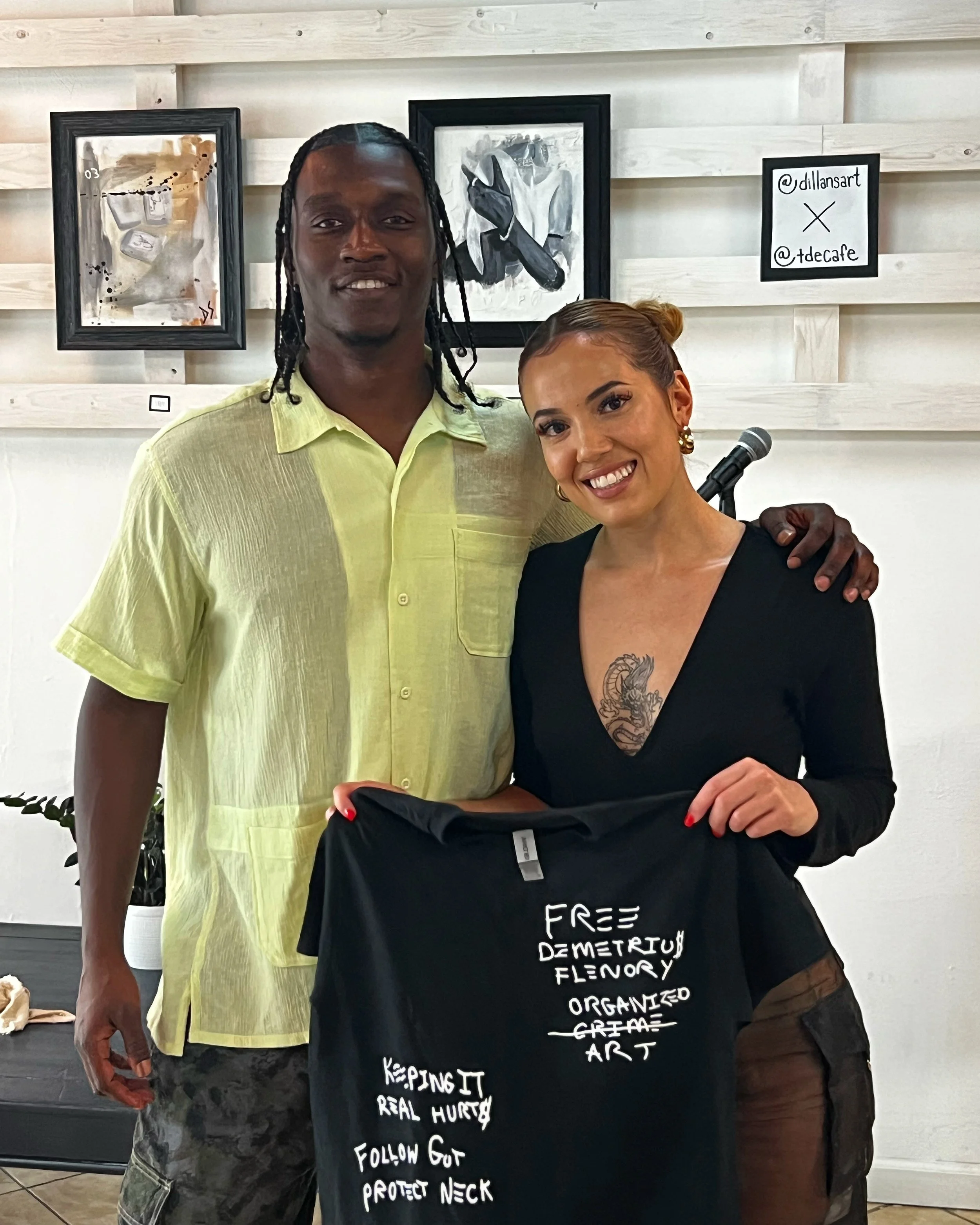 A man and woman standing together indoors, holding a black T-shirt with handwritten messages. The man, wearing a yellow shirt, has braided hair. The woman, with brown hair styled in a bun, has a tattoo on her chest and is dressed in black. The T-shirt has messages about activism and self-care, including "FREE DEMETRYU FLENORY," "ORGANIZED GRAM ART," "KEEPING IT REAL HURTS," "FOLLOW GUT," and "PROTECT NECK." In the background, there are framed artwork and a small sign with social media handles on a white wooden wall.