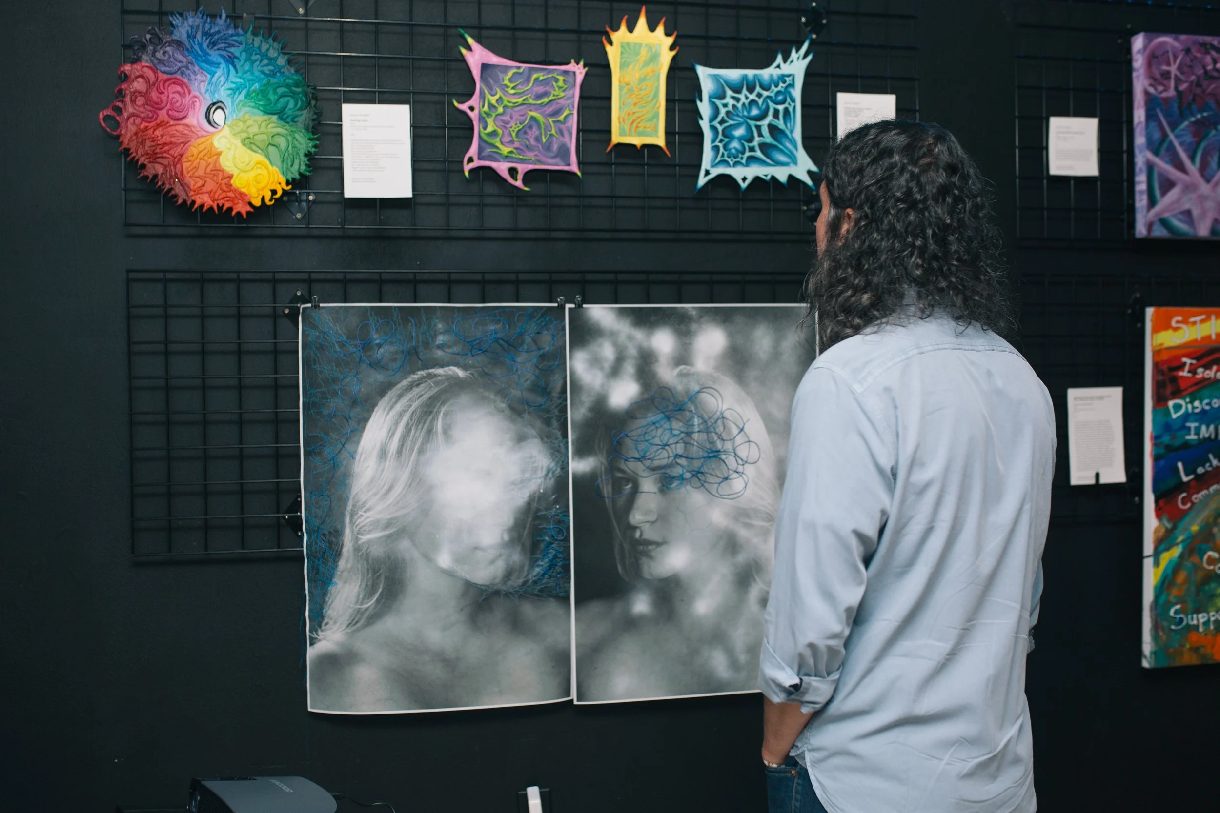 A man with long, curly hair and a light blue shirt stands in front of an art display, observing black and white photos of two women with artistic blue scribbles on their faces. The background features colorful abstract art pieces on a black wall.