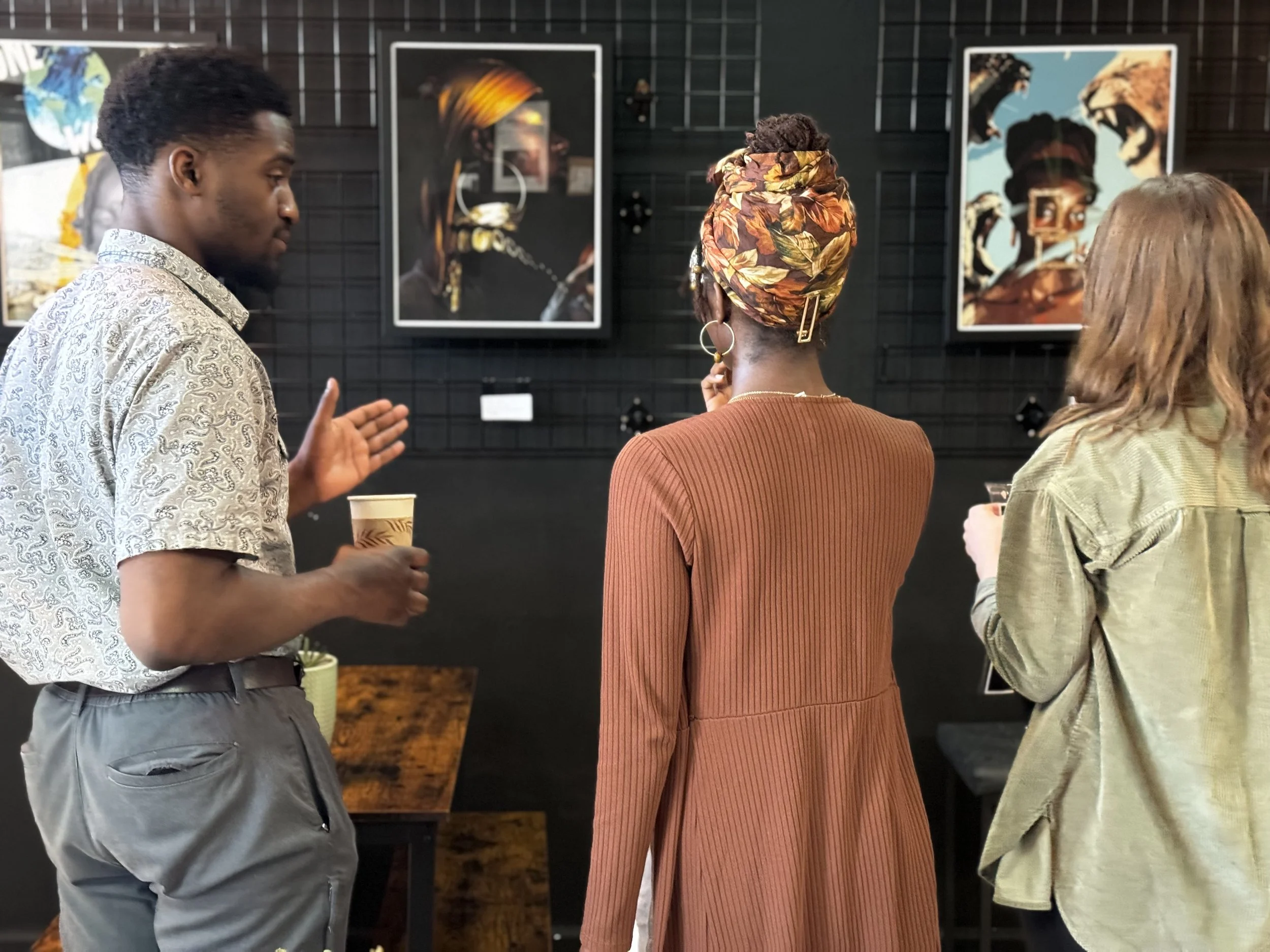 Three people are standing and talking in an art gallery with black walls and several colorful framed artworks. The person on the left is a young man with dark skin, wearing a light-colored patterned shirt and gray pants, holding a cup. The person in the center is a woman with dark skin, wearing a rust-colored dress and a colorful headscarf, with her back to the camera. The person on the right is a woman with light skin and blonde hair, wearing a light green shirt, holding a drink.