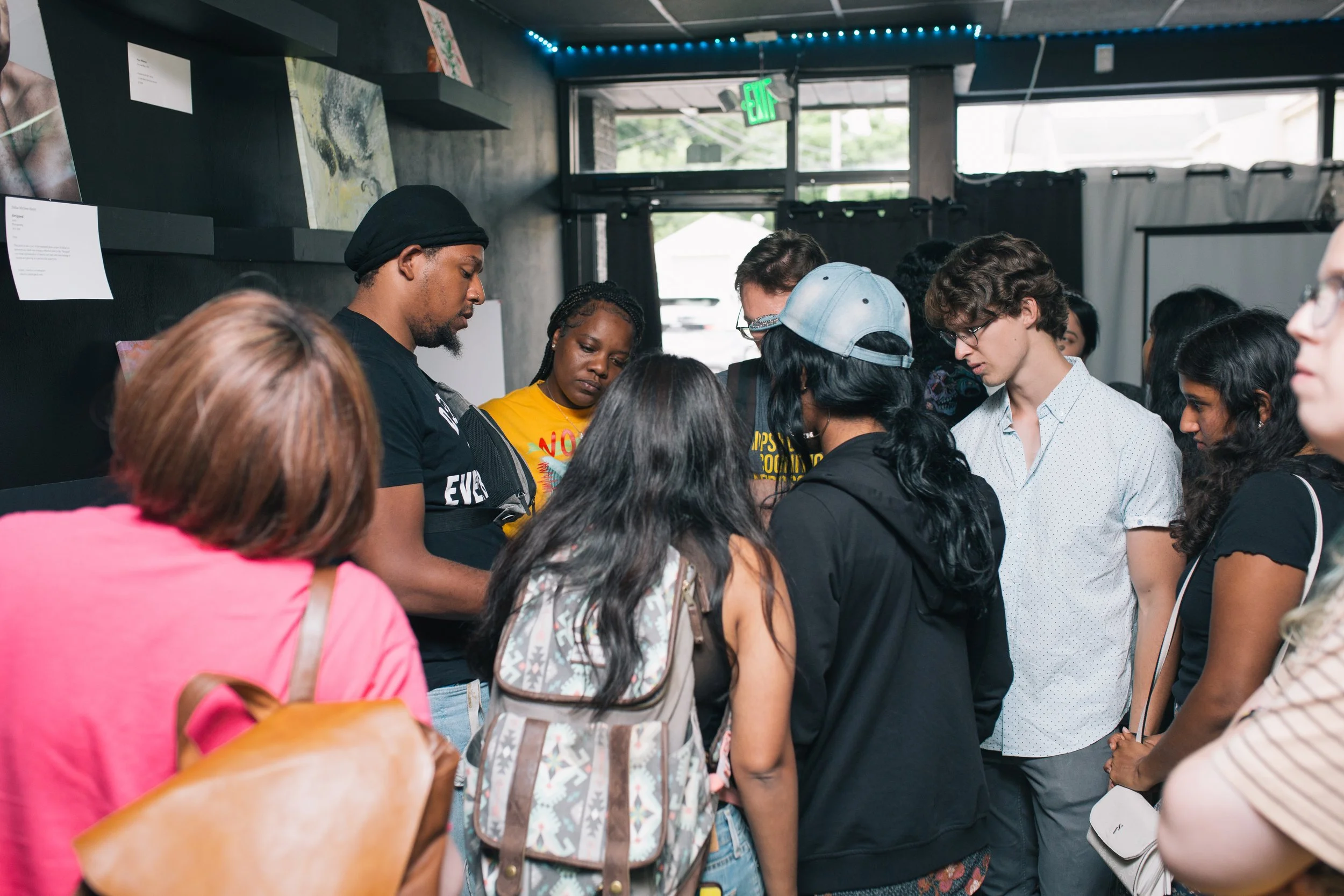 A diverse group of people gathered closely together inside a building, looking at something or listening intently, with artwork displayed on the walls and an exit sign visible in the background.