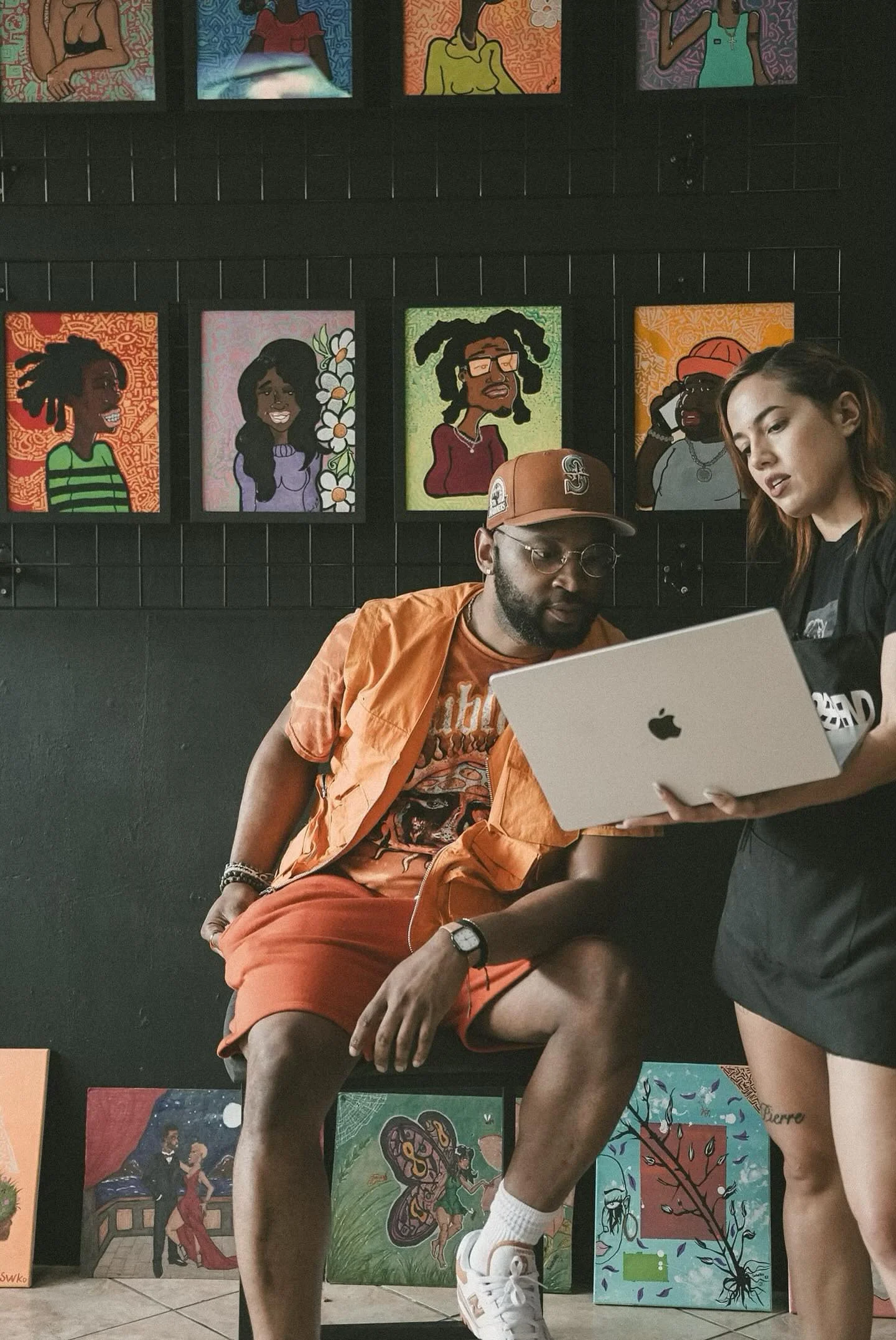 Two people looking at a laptop in front of colorful artwork depicting cartoon-style portraits hanging on a black wall.