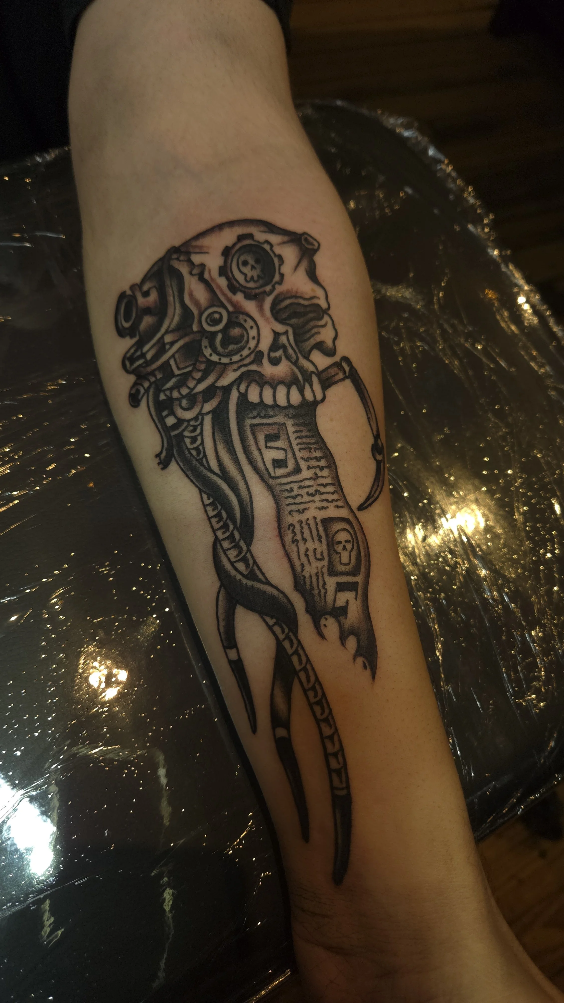 A tattoo of a servo skull from Warhammer 40,000, or Warhammer 40k. It's a skull in black and grey with machine parts implanted on it, and a long scroll hanging from it, in service of The Imperium. The Empror Protects.