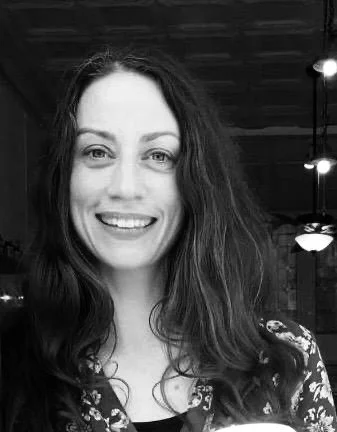 A woman with long, wavy hair smiling at the camera, in an indoor setting with ceiling lights in the background.