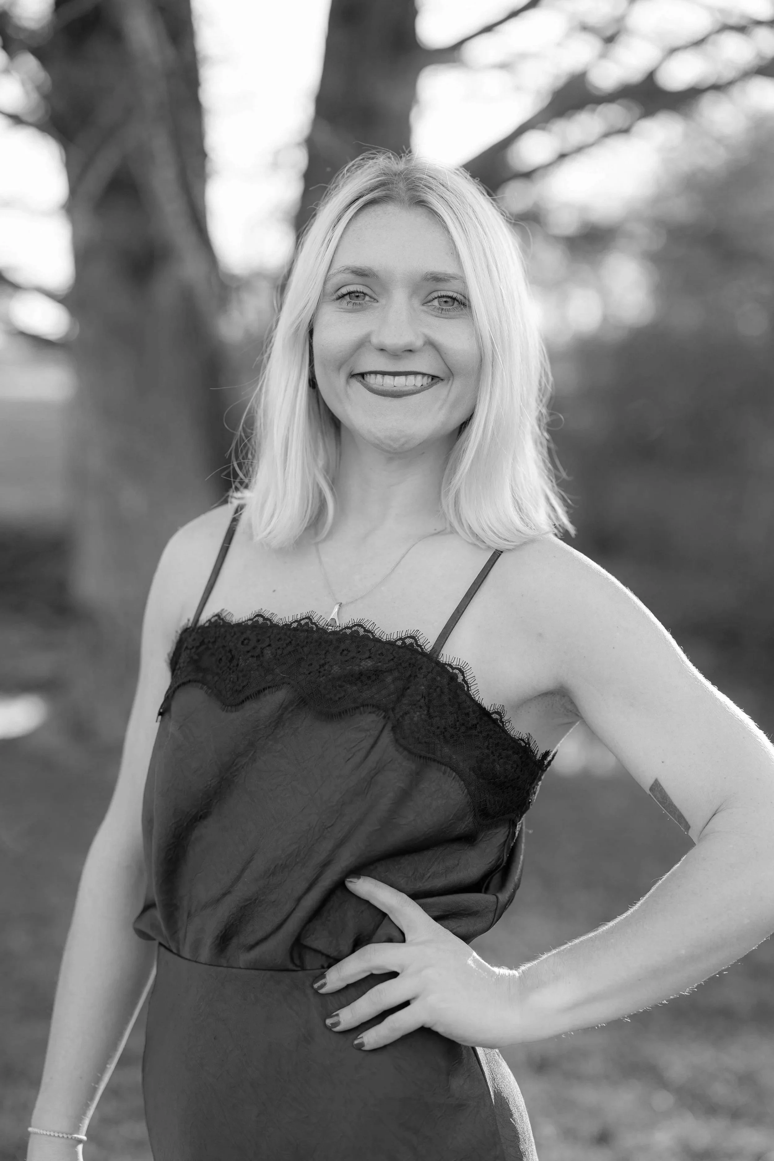 A woman with shoulder-length blonde hair smiling outdoors, wearing a dark slip dress with lace trim.