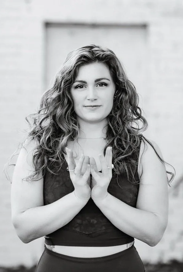A woman with long, curly hair standing outdoors, facing the camera with a neutral expression, wearing a sleeveless top and rings on her fingers.