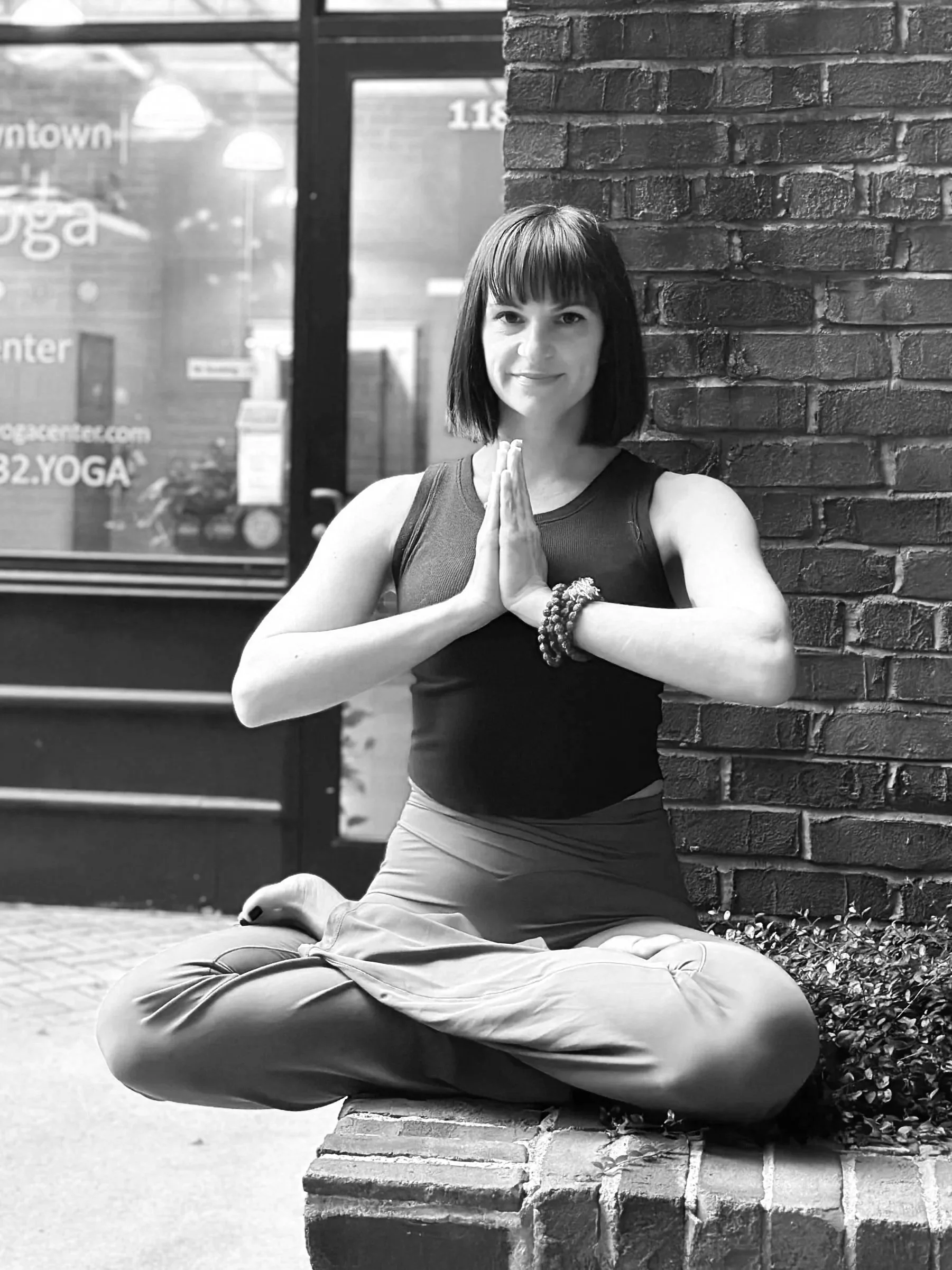 A woman practicing yoga outdoors, sitting cross-legged on a brick ledge, with hands pressed together in prayer position, smiling at the camera.