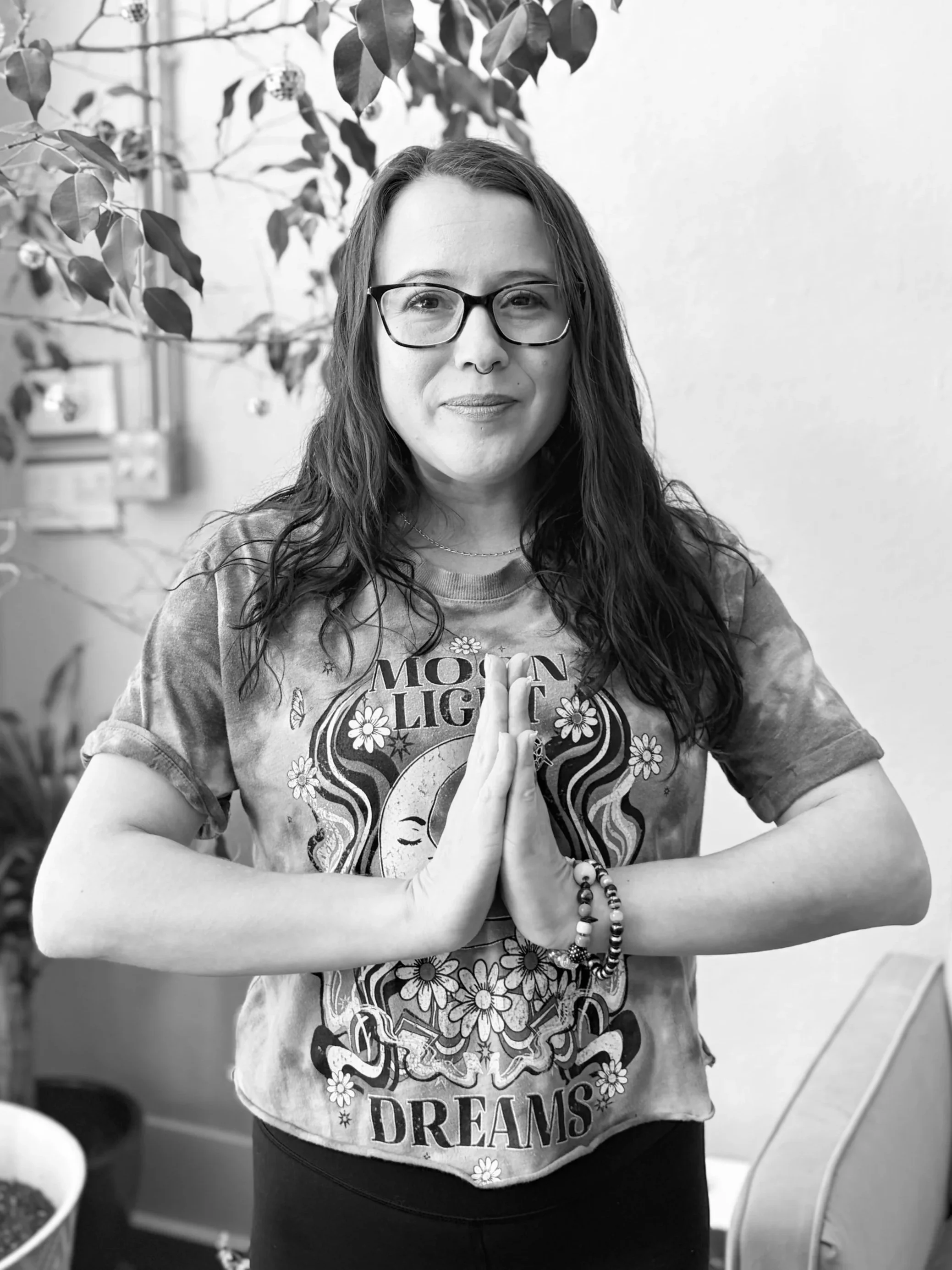 A woman with glasses and long, wavy hair standing indoors with plants in the background. She is wearing a tie-dye t-shirt with a moon, flowers, and the words 'Moon Light Dreams.' She has her hands pressed together in a prayer or greeting gesture, wearing bracelets on her wrist.