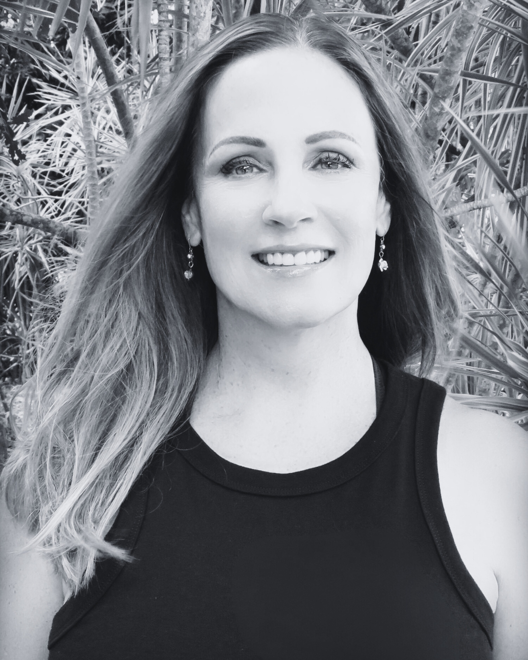 A woman with long hair smiling outdoors, wearing earrings and a sleeveless top, with plants in the background.
