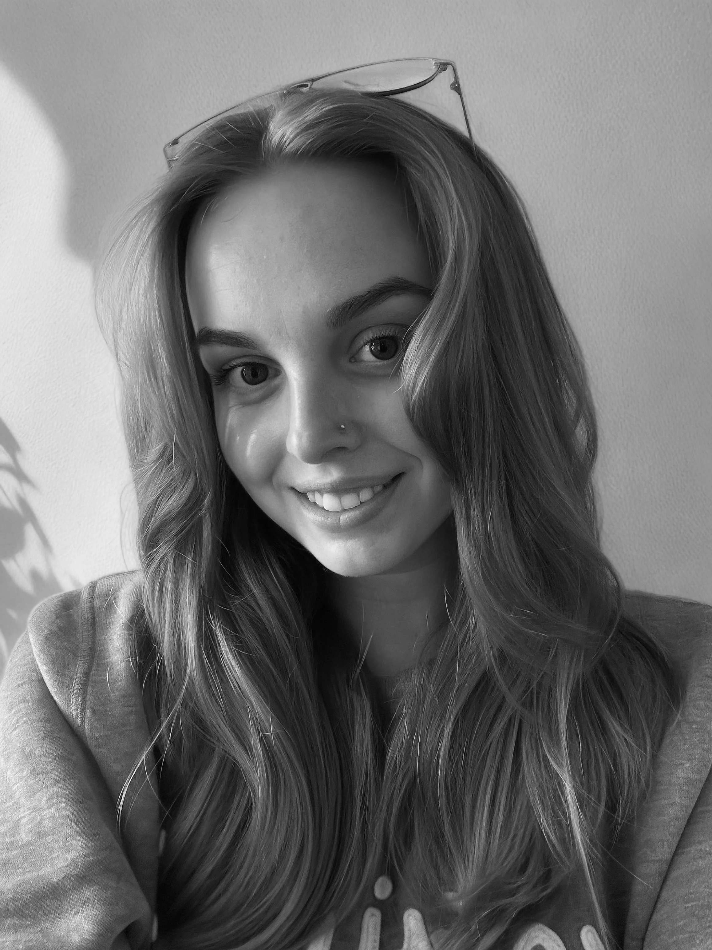 Black and white photo of a smiling woman with long, wavy hair, wearing glasses on her head and a nose piercing, looking at the camera.
