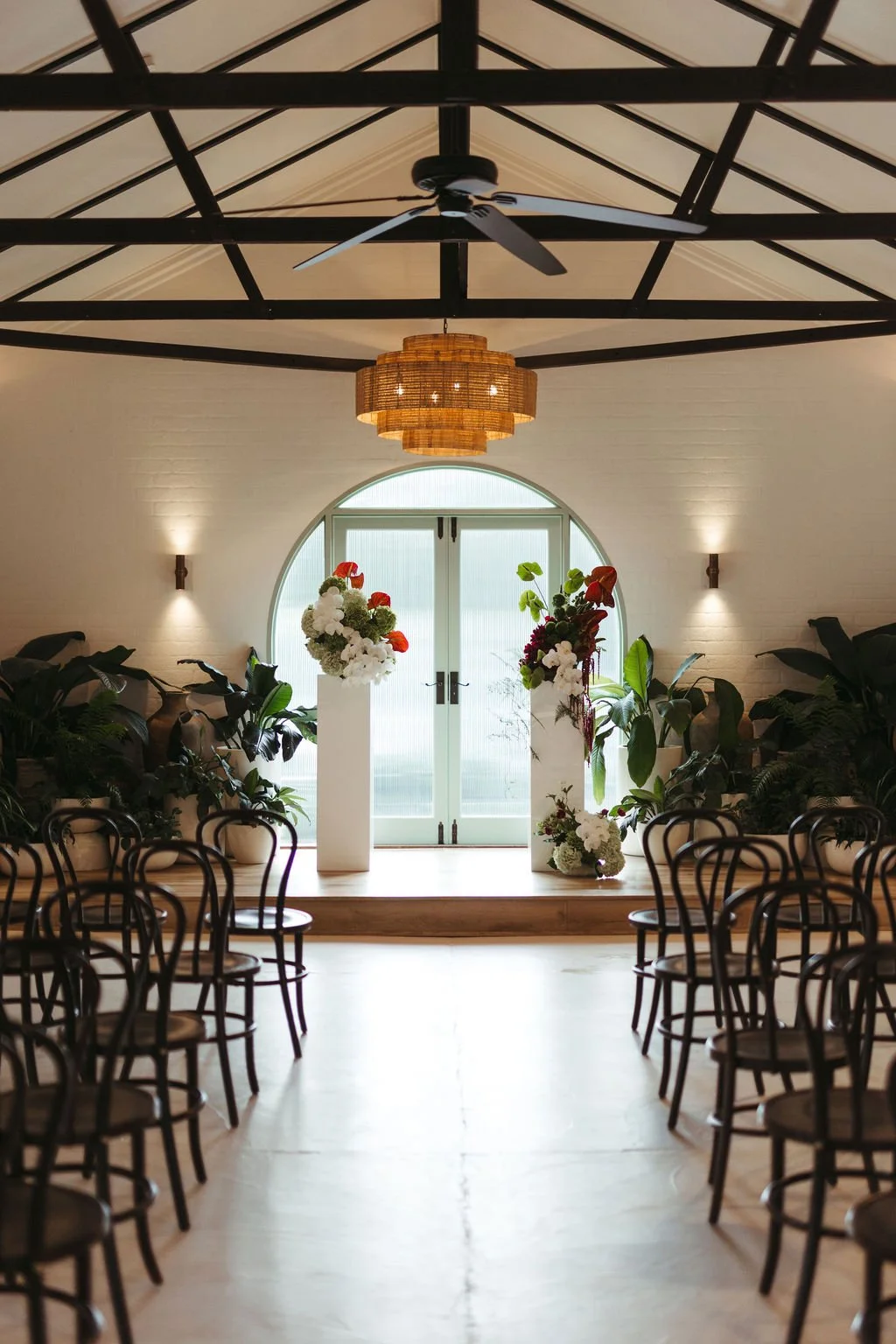 An elegantly decorated indoor space with rows of chairs on either side of a central aisle, leading to an arched window and double doors. The space features a high ceiling with exposed black beams, a wicker chandelier, and contemporary wall-mounted li
