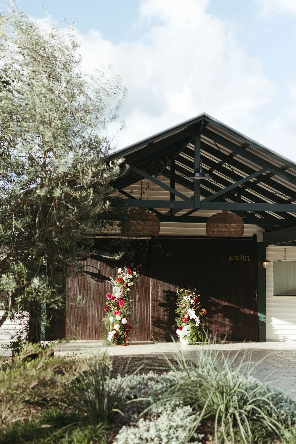 The image shows the entrance of a building with a dark wooden door, decorated with floral arrangements on either side. The building has a metal roof with two hanging woven lamps, and the surrounding area includes greenery and a partly cloudy sky.