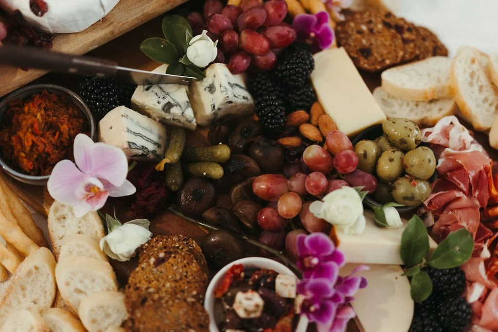 A cheese and fruit platter with grapes, blackberries, olives, prosciutto, and crackers, decorated with purple and white orchids.