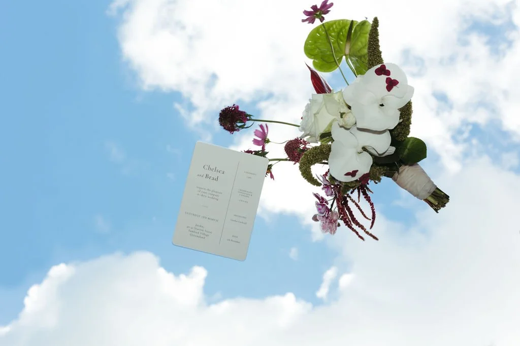 A wedding bouquet with white orchids, purple and pink flowers, and greenery, attached to a card that reads Chelsea and Brad, against a partly cloudy blue sky.