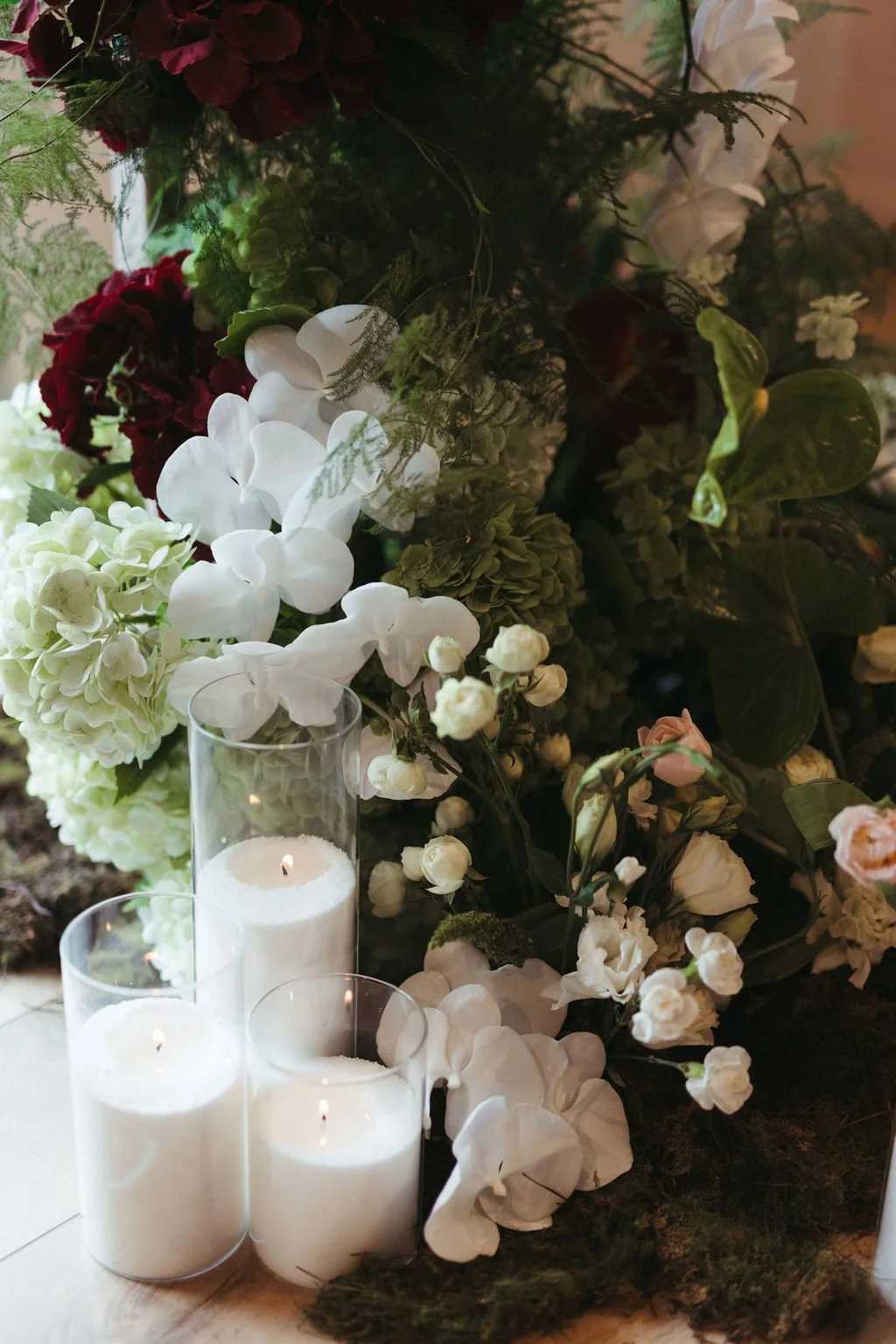 A floral arrangement with white, red, and pink flowers, surrounded by green leaves, and lit white candles in glass holders.
