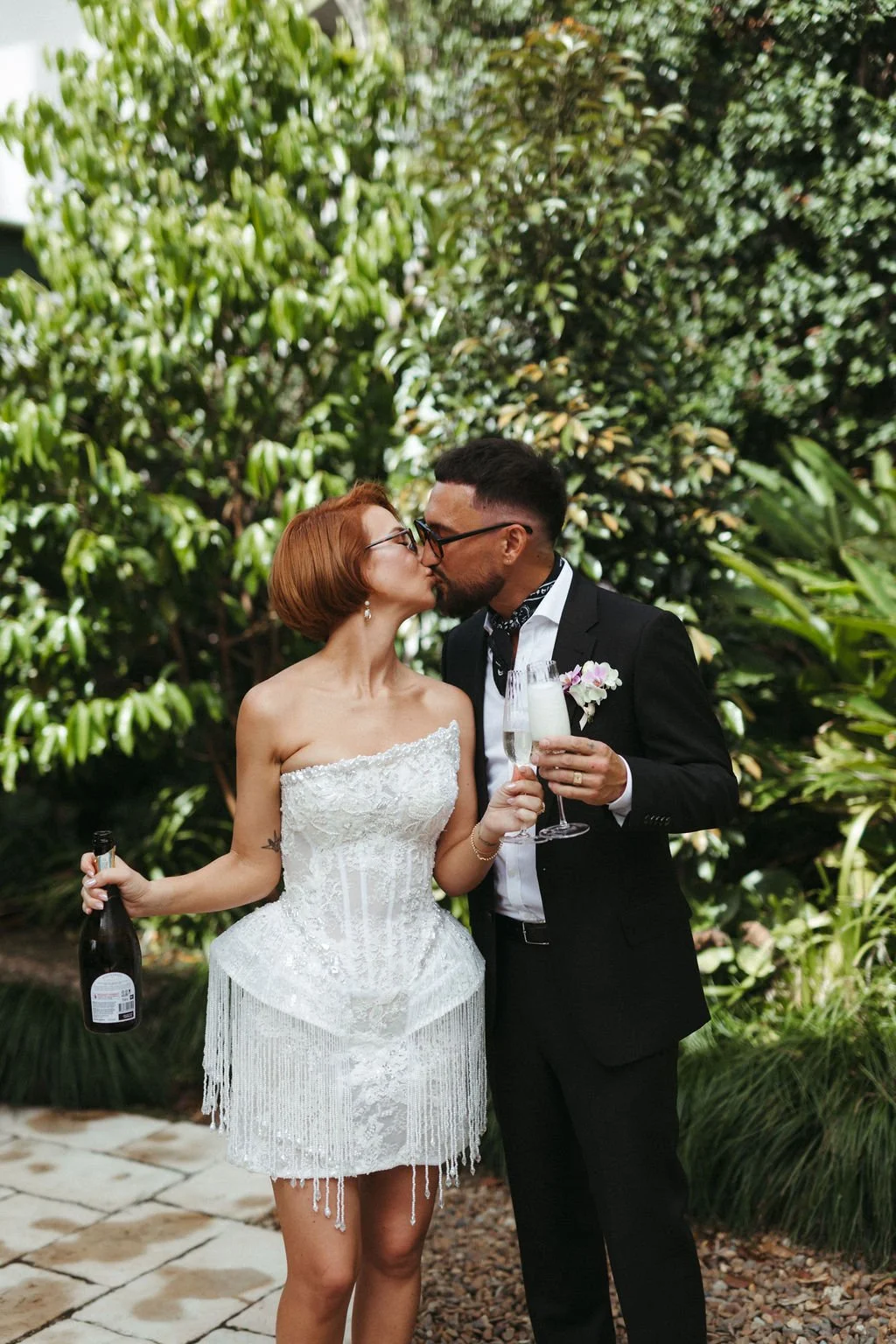 A newlywed couple sharing a kiss outdoors; the bride in a strapless white lace dress holding a bottle of champagne, the groom in a black tuxedo with a flower boutonniere and a glass of champagne.