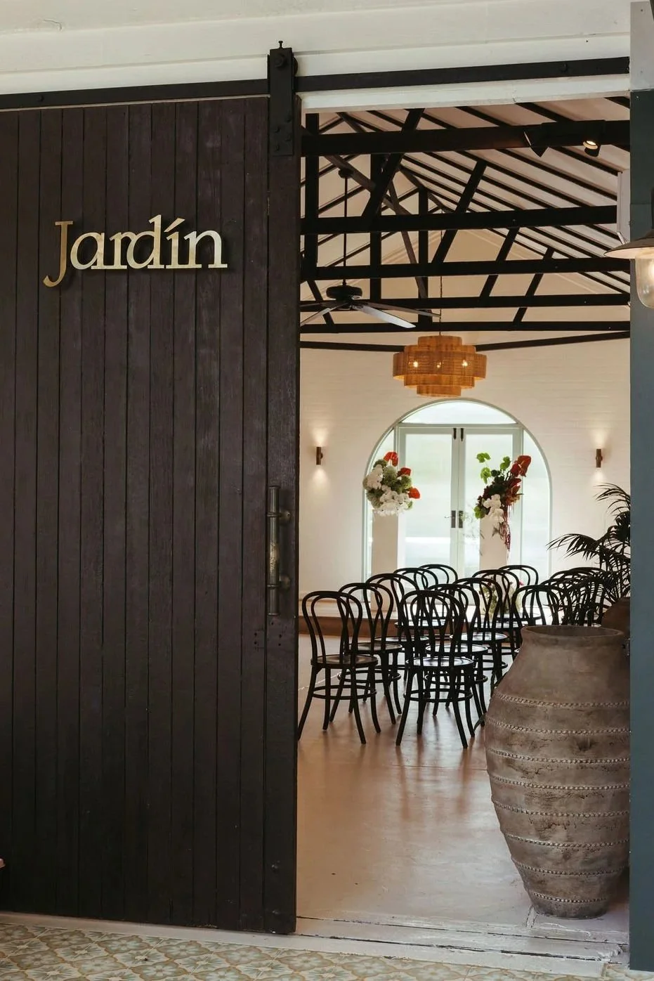 Stylish entrance door with the word 'Jardin' and view of a dining area with black chairs, large flower arrangements, and a pendant light