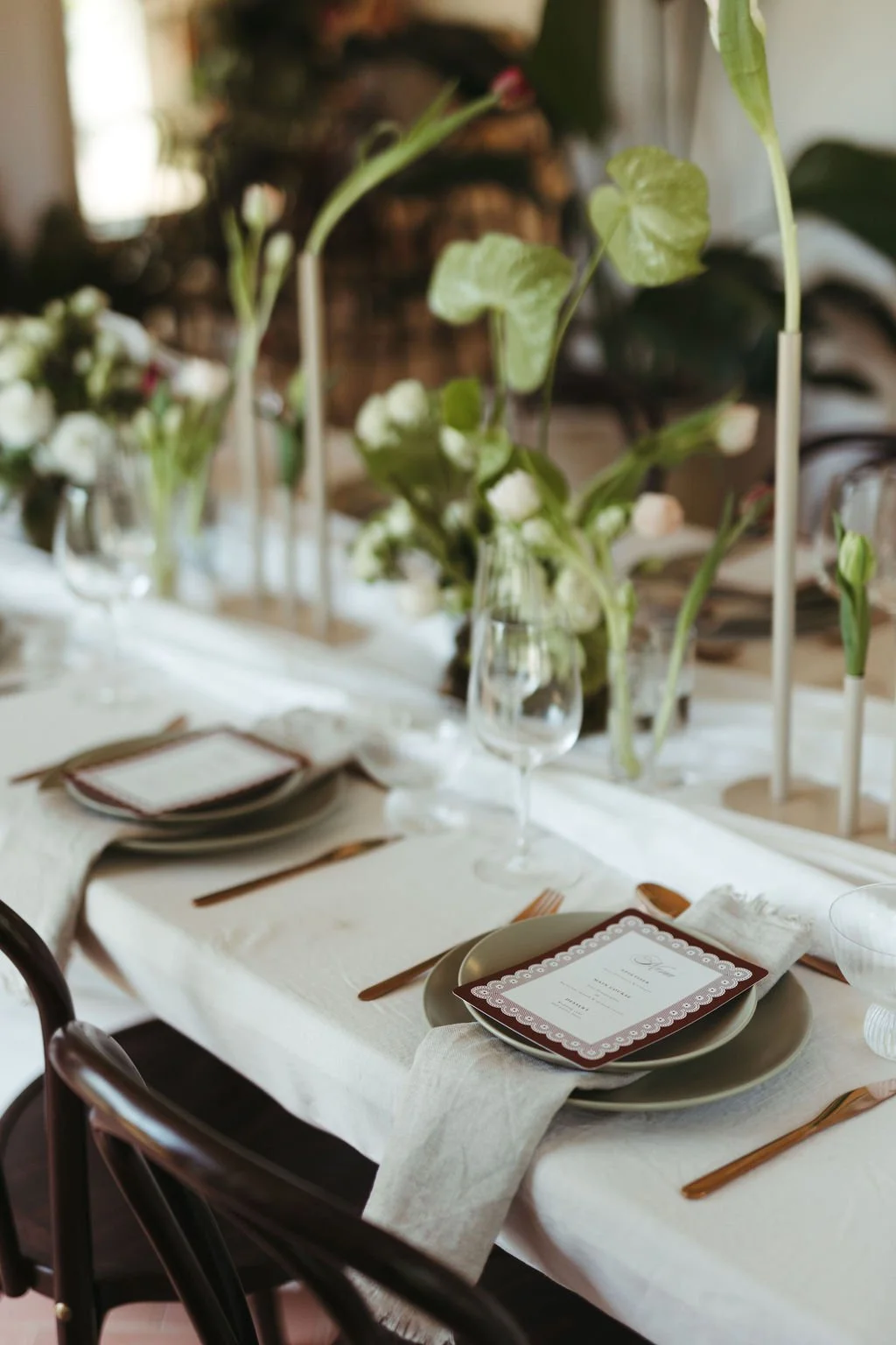 A long dining table set for an event with white tablecloth, green plates, wooden utensils, wine glasses, and a menu card. Tall vases with green leafy stems and white flowers decorate the center.