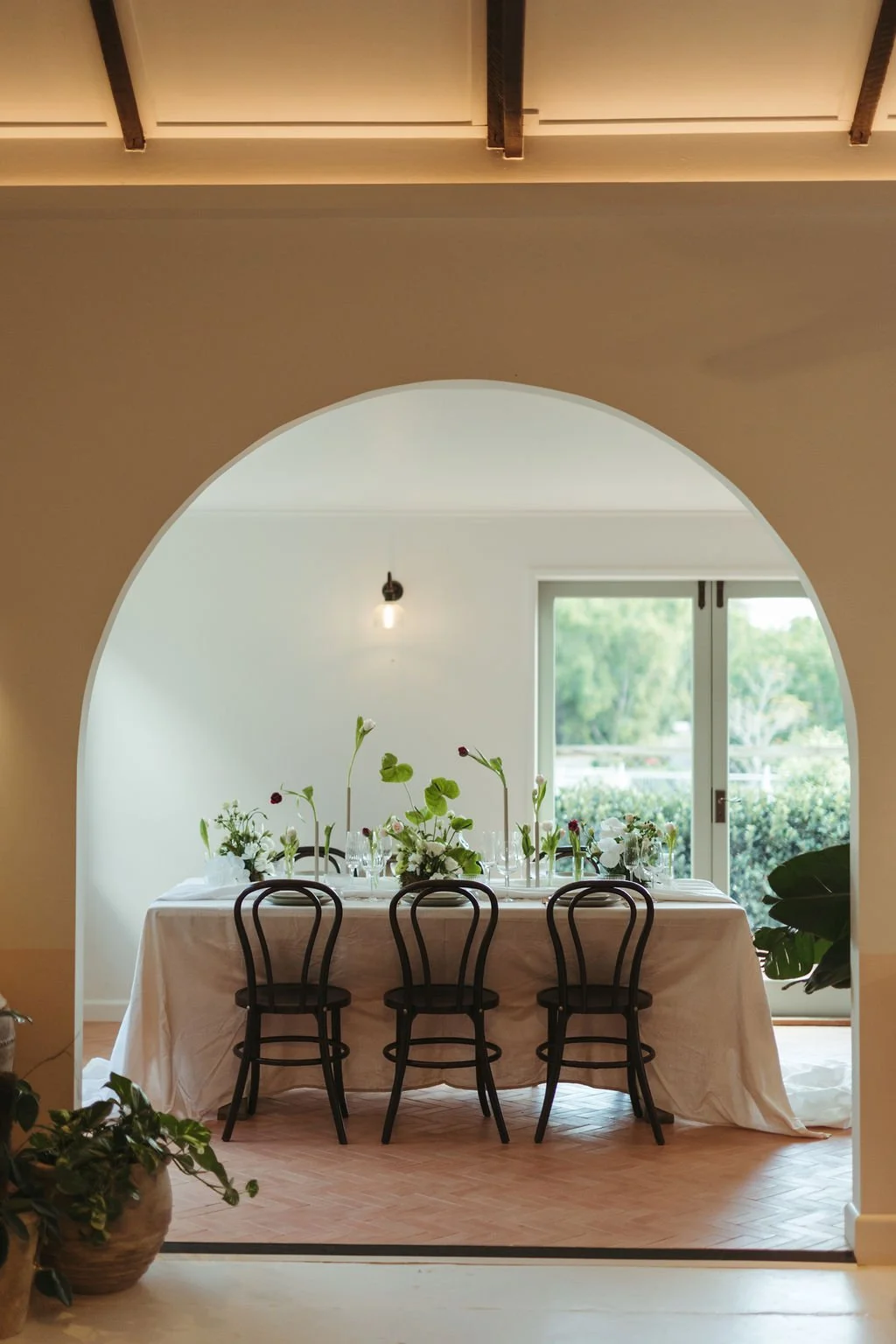A dining table set with white tablecloth, floral arrangements, and glassware, viewed through an arched doorway in a bright room with large windows and greenery outside.