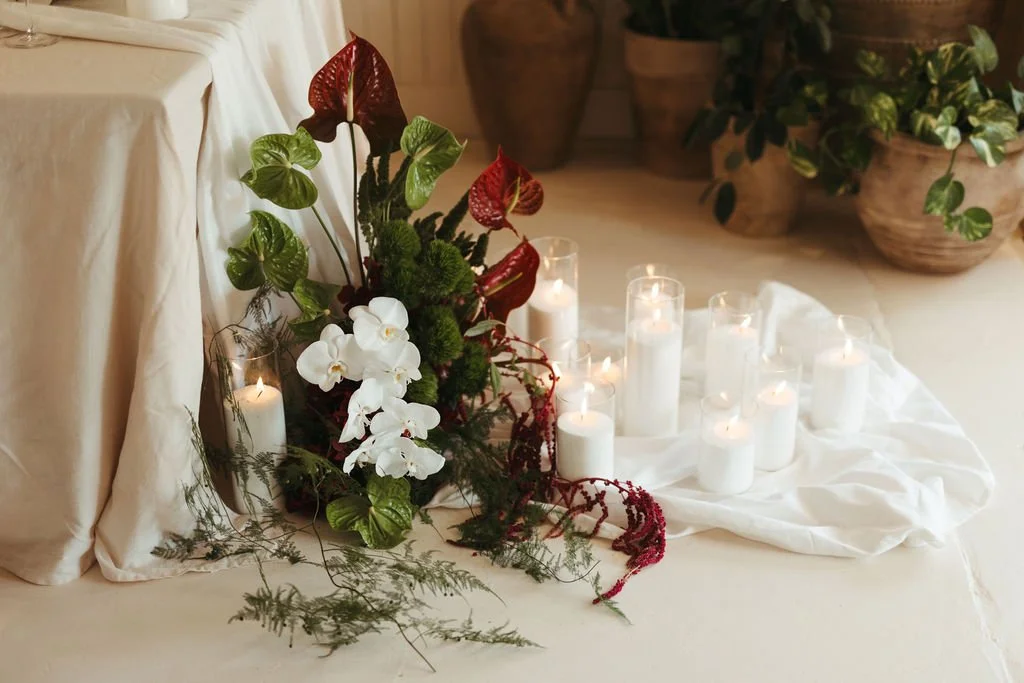 A decorative arrangement featuring white candles, red and green plants, white orchids, and greenery on a cream-colored surface, with additional potted plants in the background.