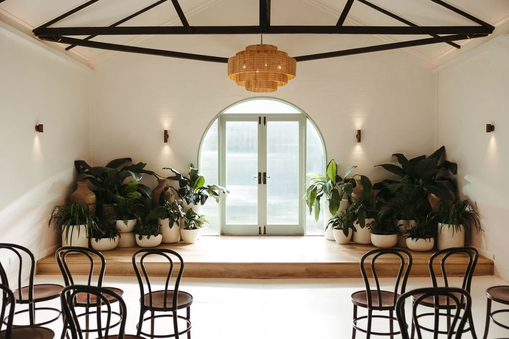 Interior of a bright, minimalist room with a small stage, potted plants, and chairs facing the stage.