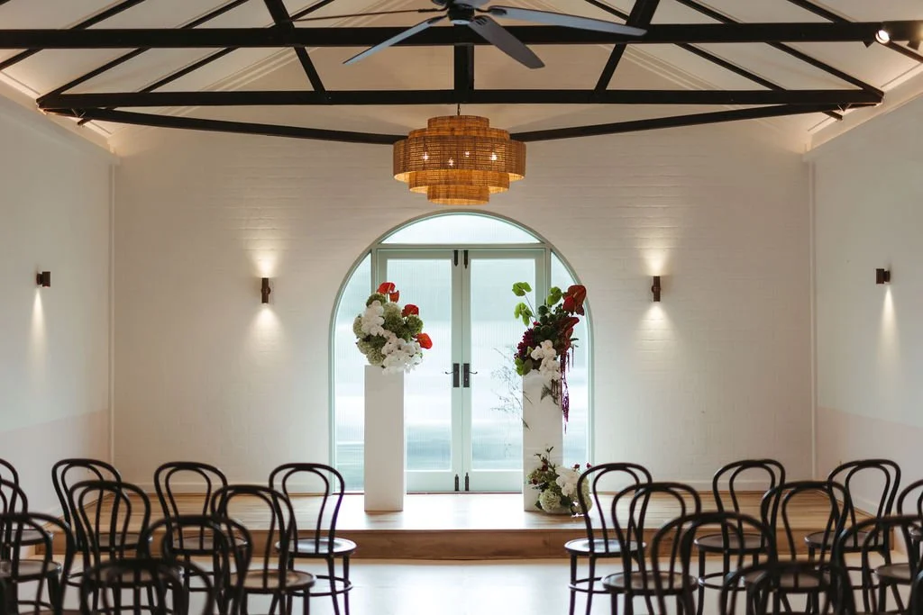 Indoor event space with rows of black chairs facing a small stage with floral arrangements, a back door, and a ceiling fan.