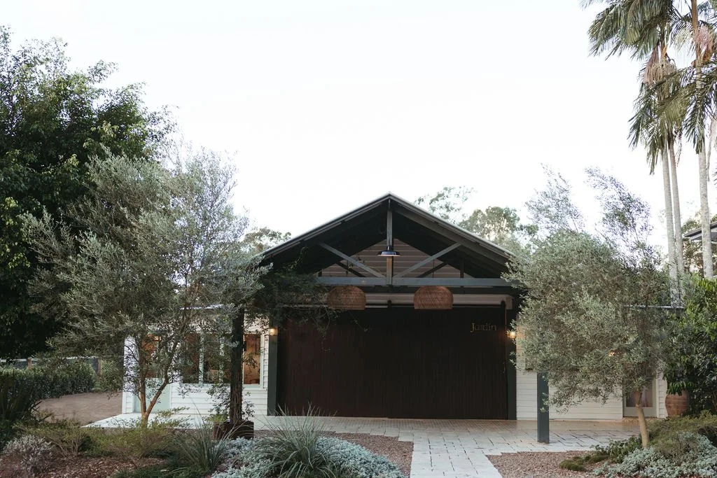 A modern house with a dark wooden garage door, surrounded by trees and shrubs, with a paved walkway leading to the entrance, and a ceiling fan hanging under the roof over the driveway.