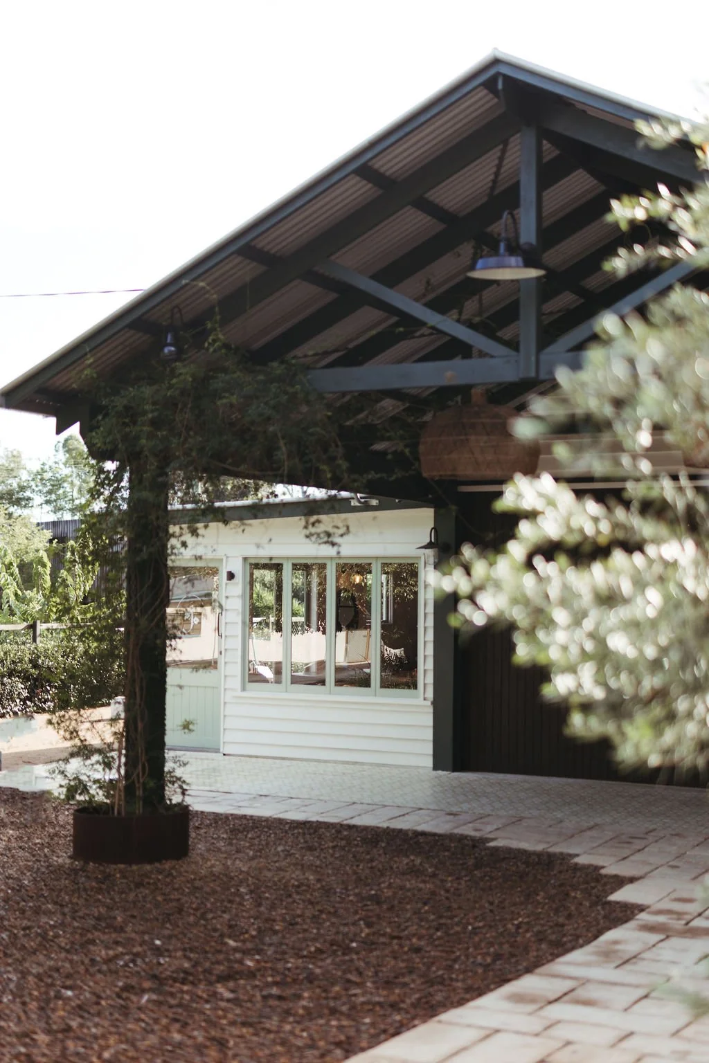 A modern wedding event venue with a covered porch, white siding, large windows, and an overhanging roof with dark beams, surrounded by trees and a landscaped yard.