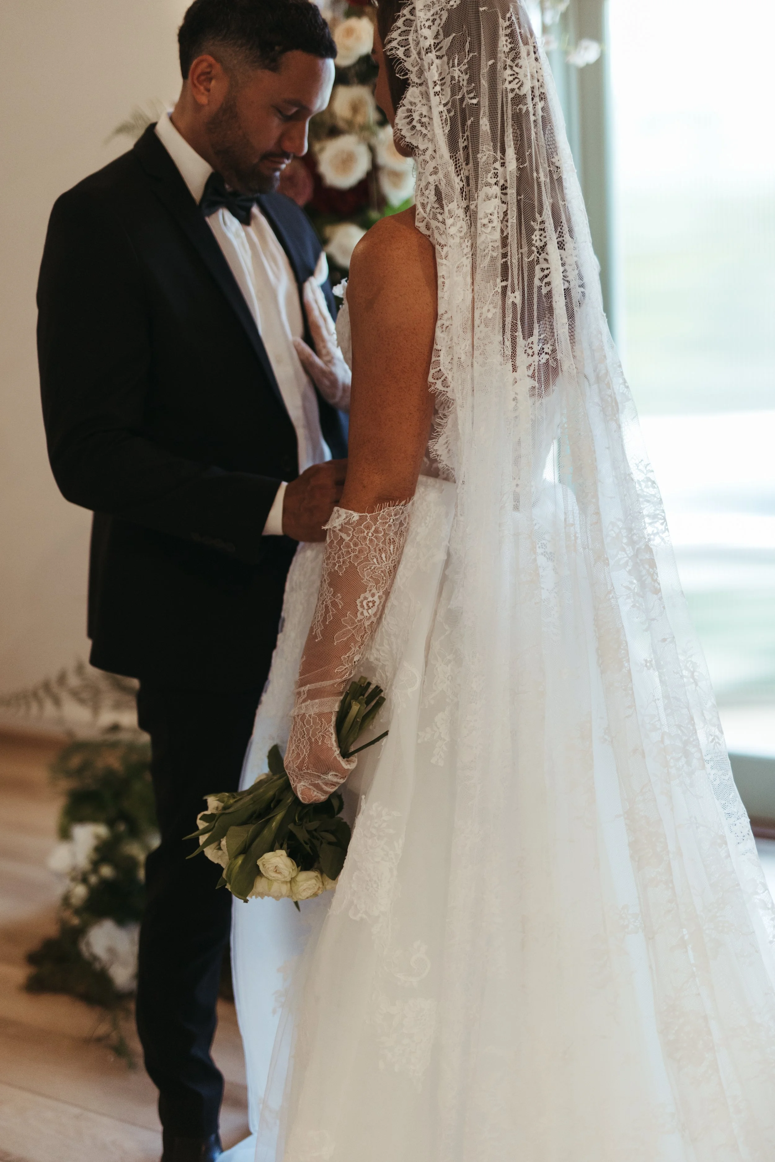 A bride and groom kneeling with heads bowed and eyes closed. The groom wears a black tuxedo with a bow tie, and the bride is in a white lace wedding gown with a lace veil. The bride holds a bouquet of white roses. The background features soft natural