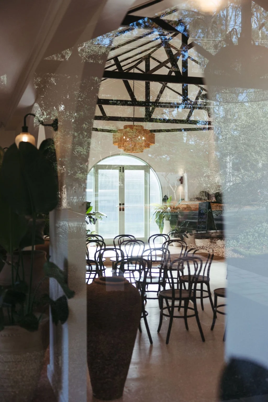 Reflection of a room with black chairs, large arched window, green plants, and a chandelier, seen through a glass door or window with a double exposure effect of trees and sky outside.