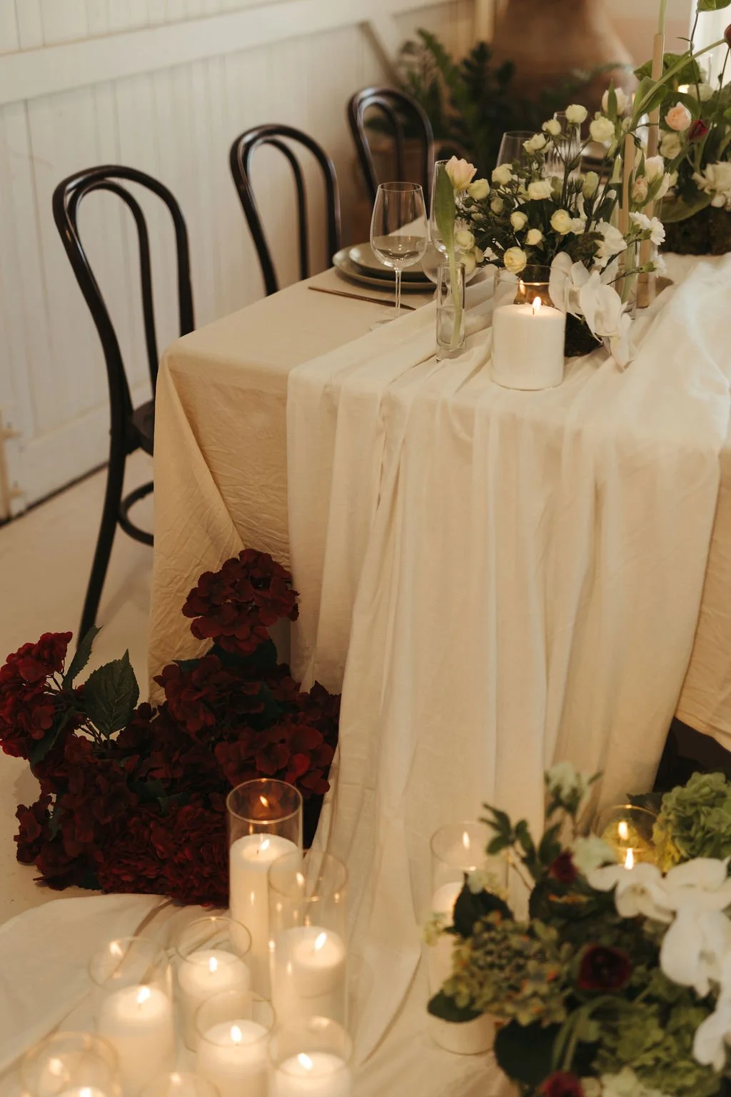 A decorated table with white tablecloth, floral arrangements, candles, and empty glassware, set in a cozy indoor setting.